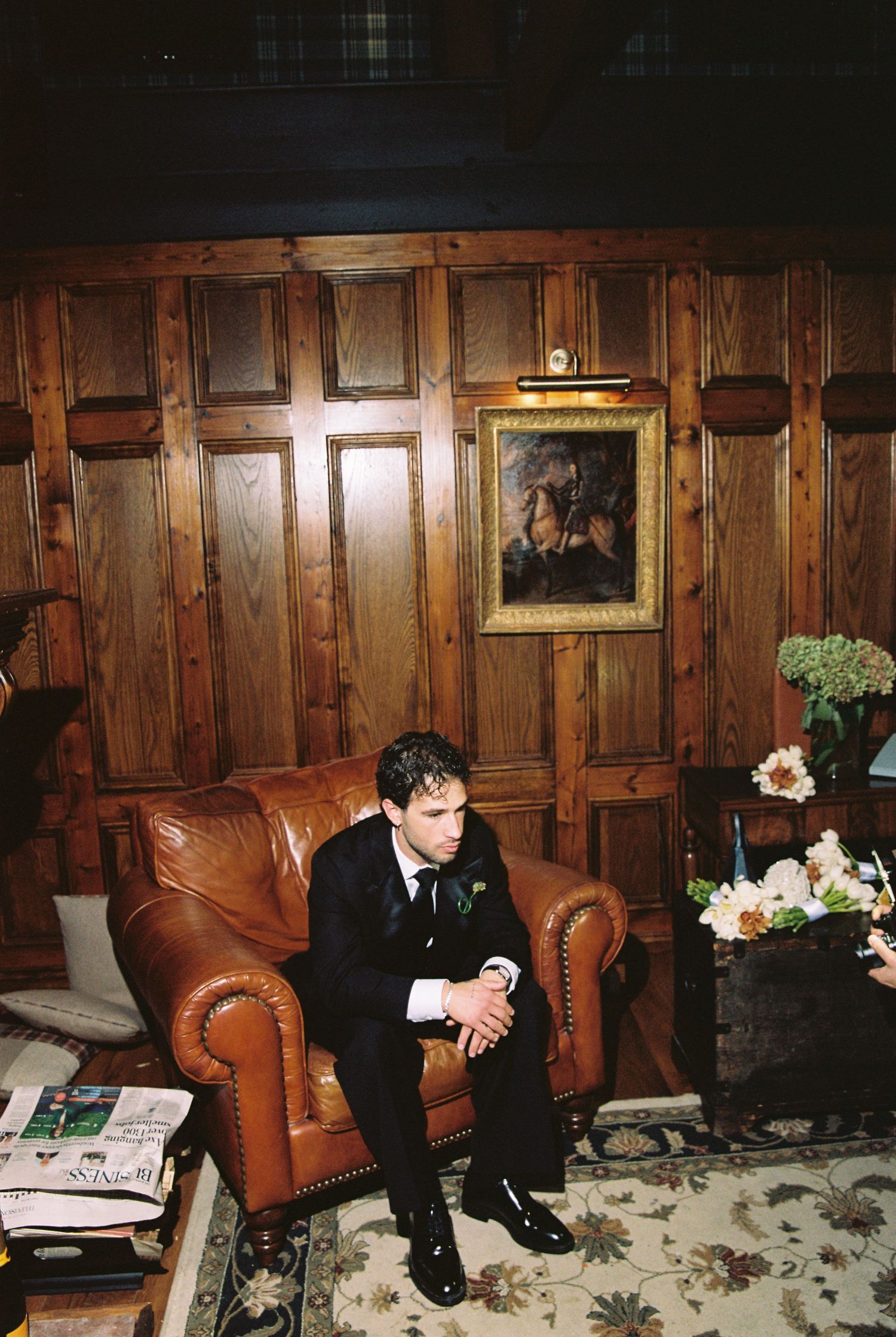 Groom in black suit sitting alone on leather armchair in elegant wood-paneled room.