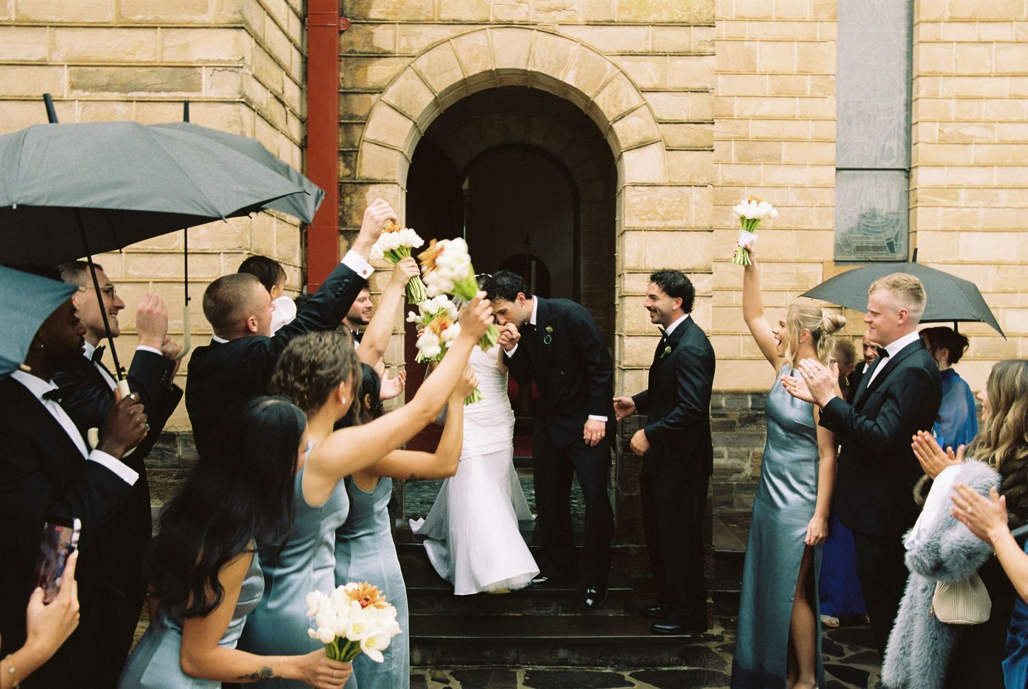 Newlywed couple exits church amid cheering wedding guests holding bouquets and umbrellas on a rainy day.