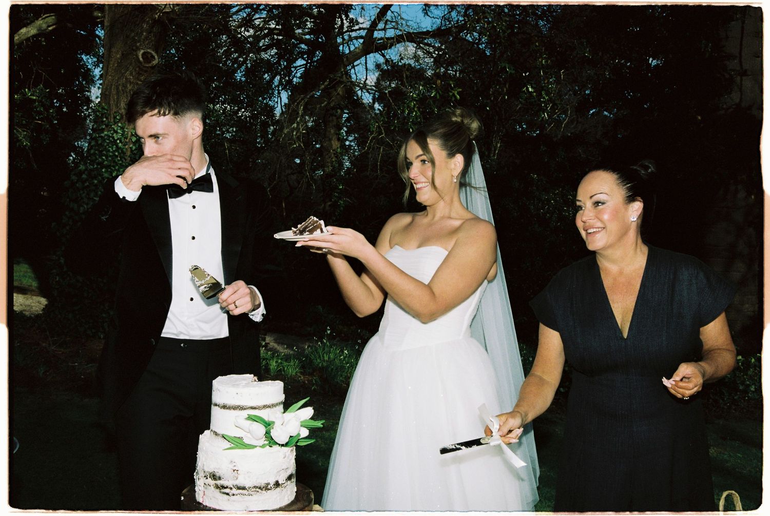 Bride and groom cut wedding cake outdoors while female guest smiles nearby at evening reception.