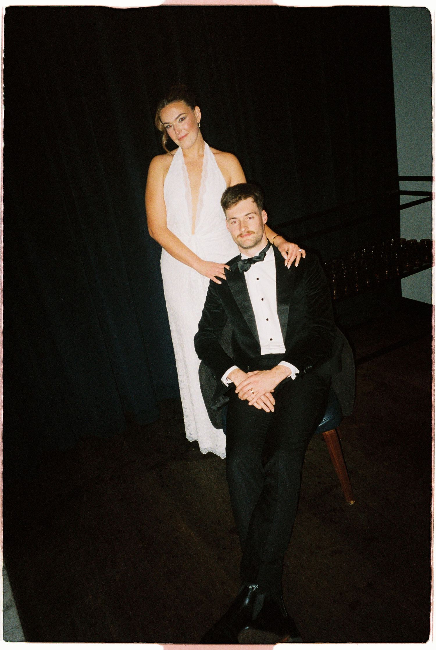 A bride in a white halter gown stands behind a seated groom in a black tuxedo against a dark curtain backdrop.