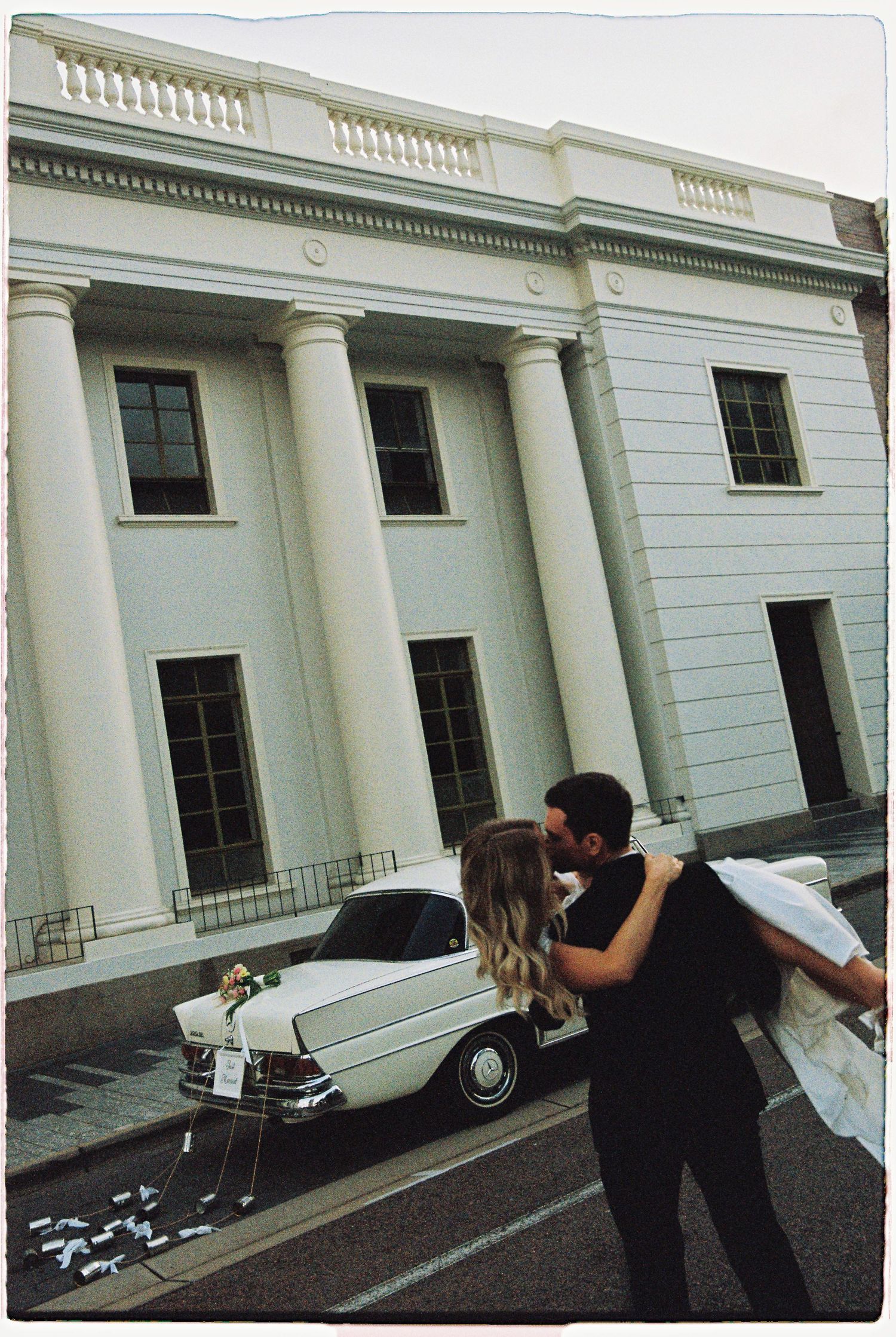 Groom lifts bride in kiss outside grand white neoclassical building with vintage Mercedes wedding car.