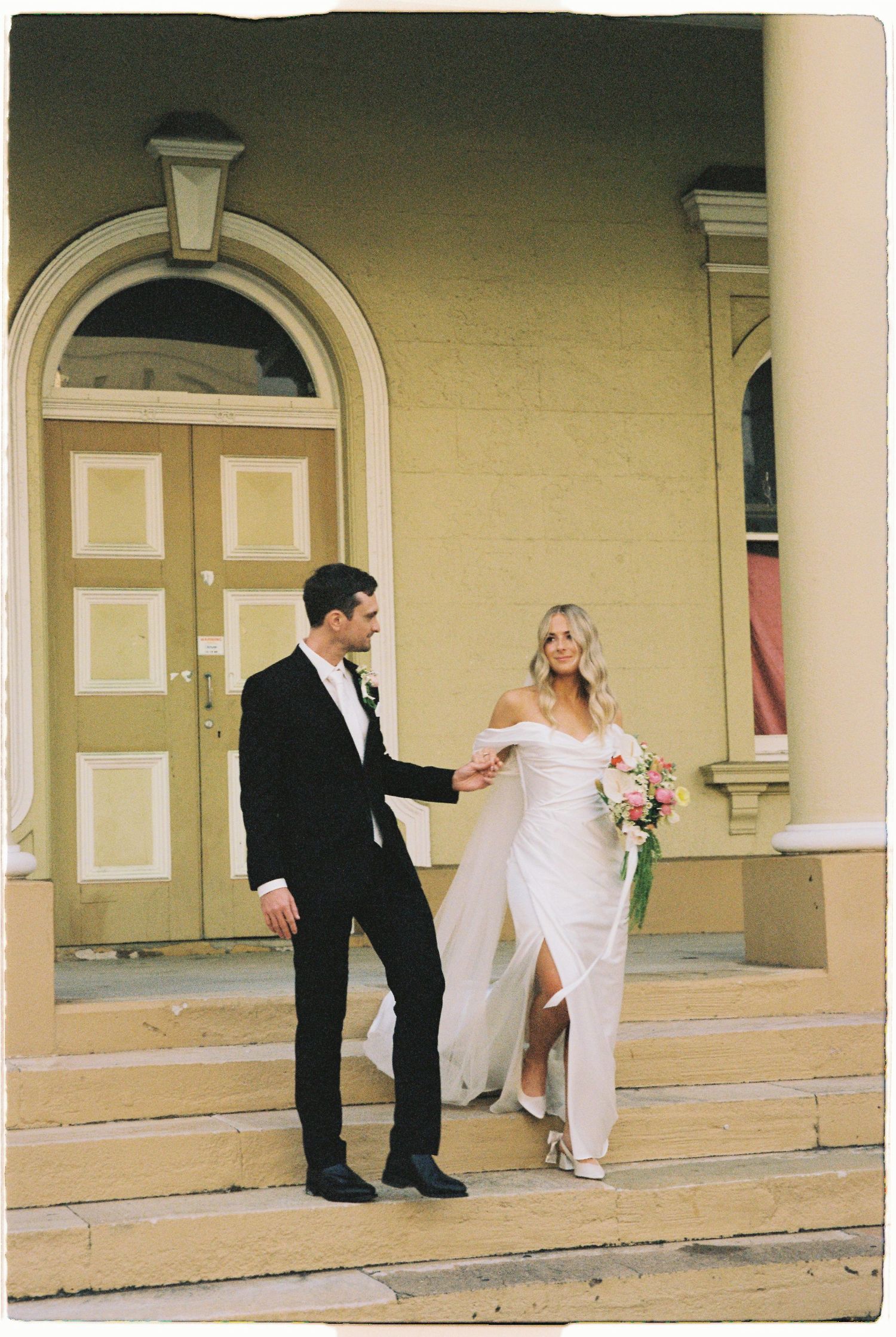 Bride in off-shoulder white gown holds groom's hand on church steps, both smiling, with a pink bouquet.