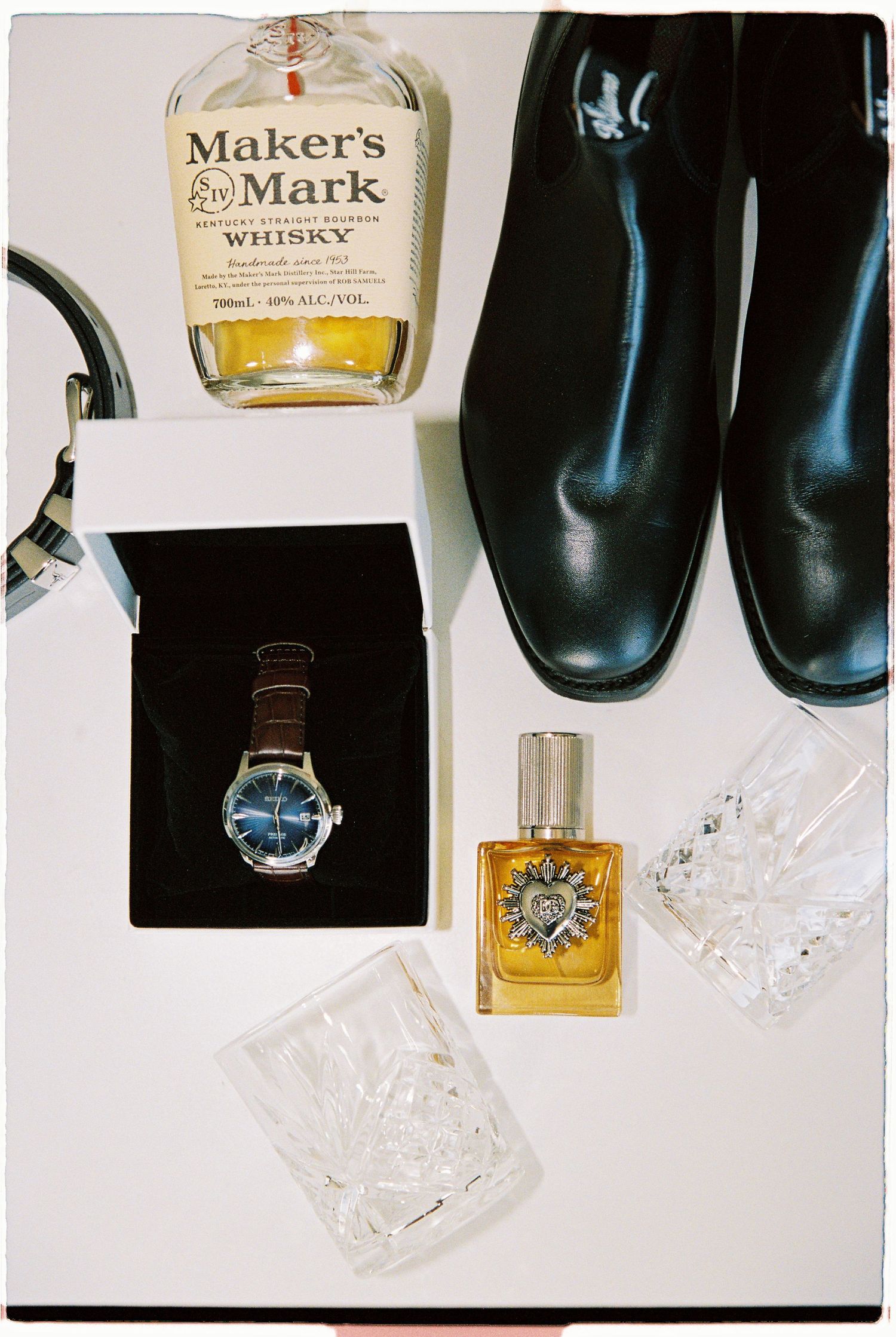 Flat lay of groom accessories including Makers Mark whisky, Seiko watch, cologne, black shoes, and crystal glasses.