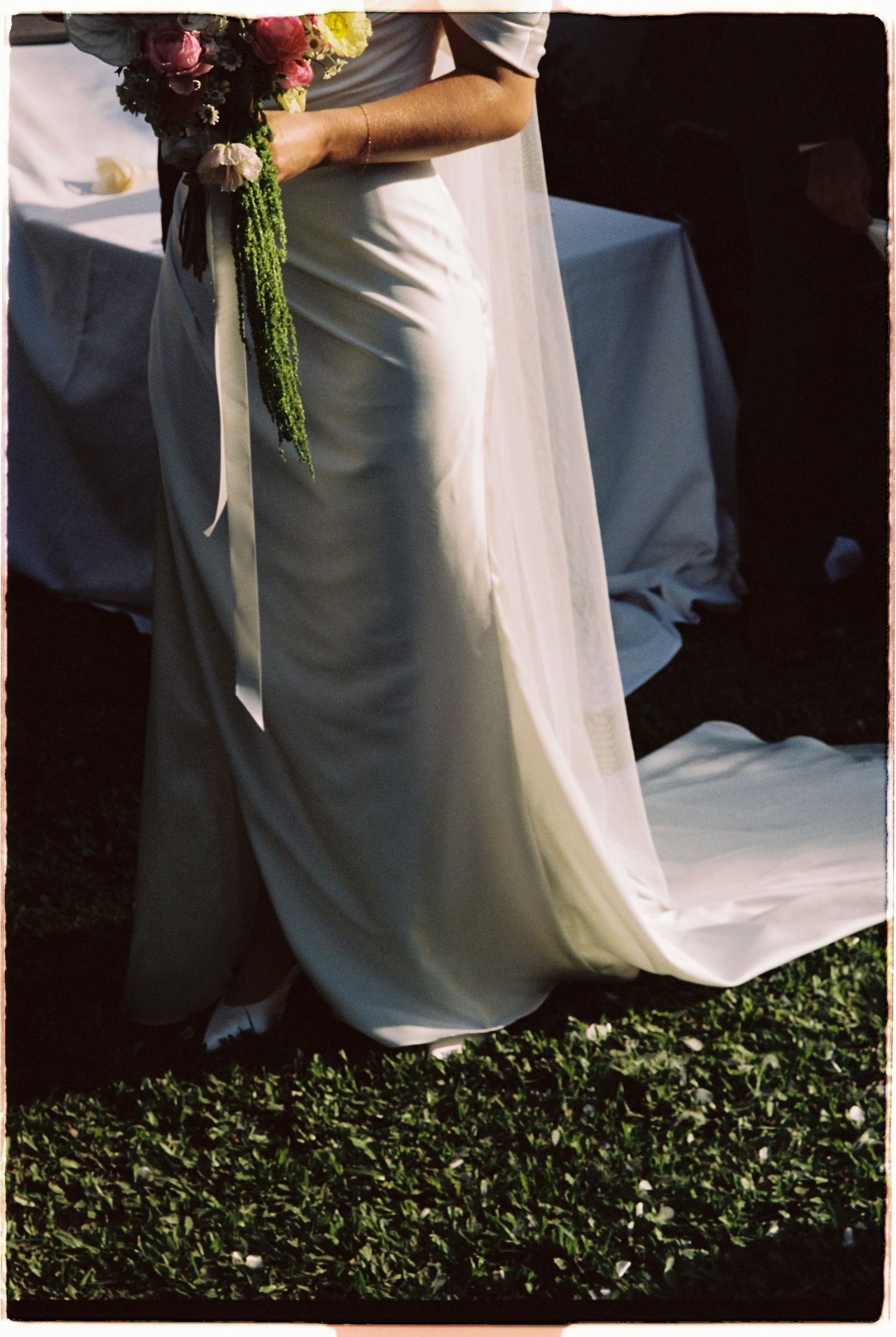 Bride in sleek white satin gown holding vibrant bouquet with long flowing veil on grass.
