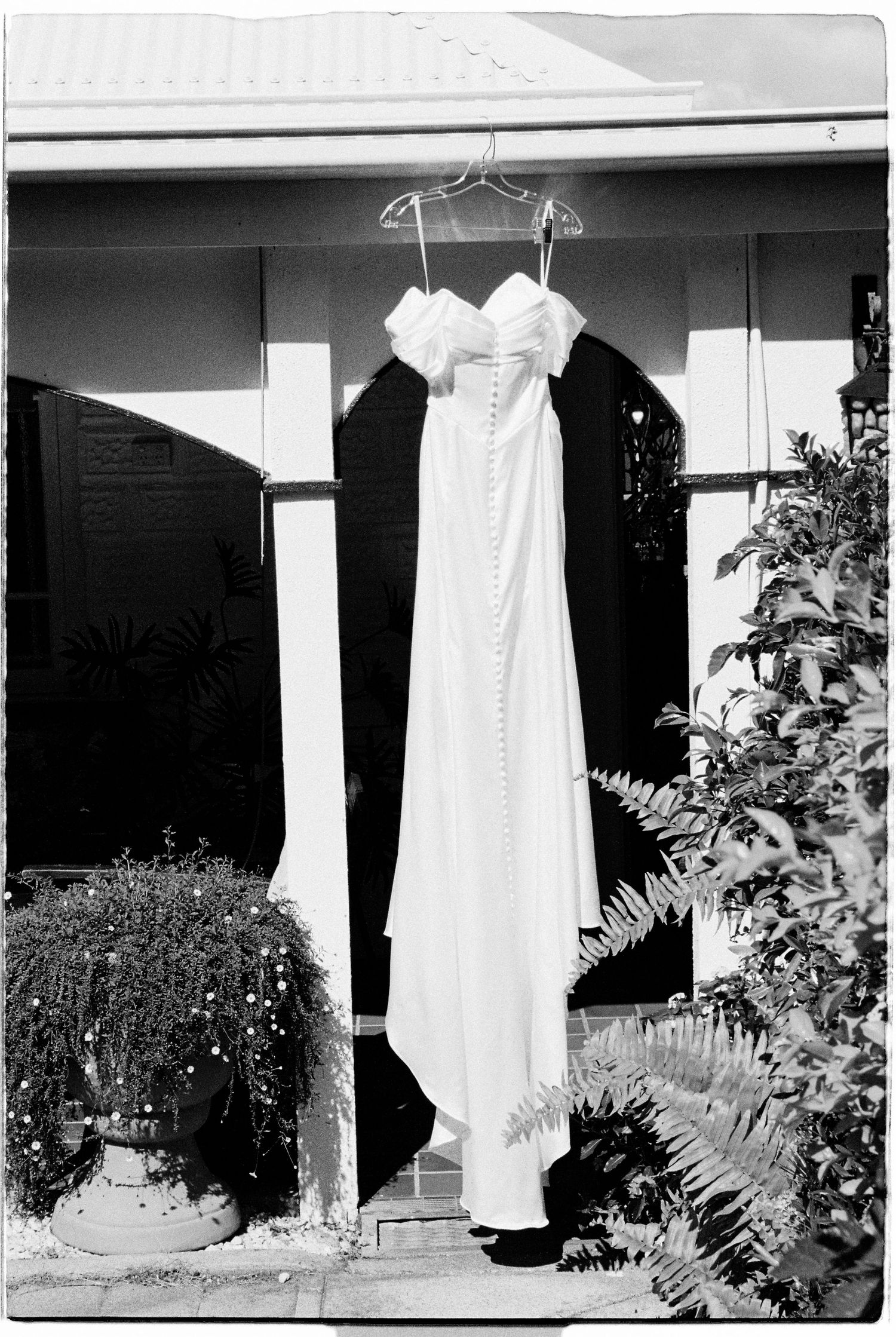 White off-shoulder wedding dress with button back hanging on hanger outside a house, black and white.