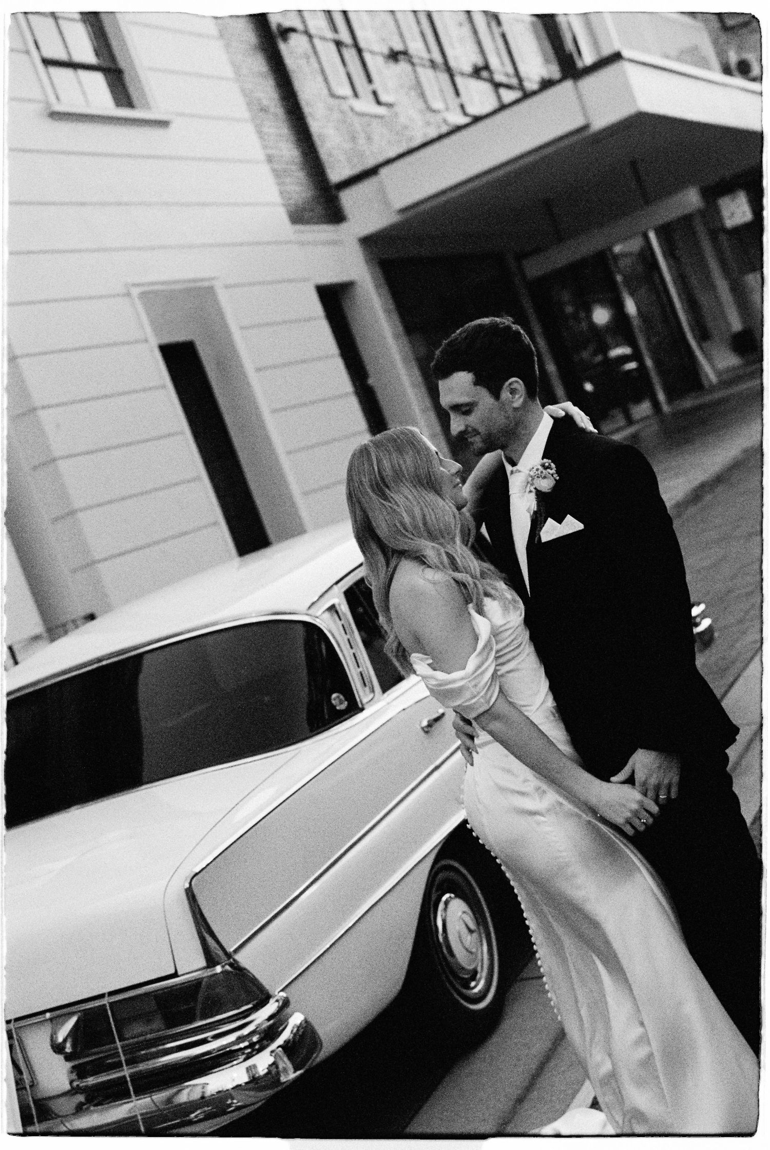 Black and white photo of bride and groom sharing a kiss beside a vintage white Mercedes.