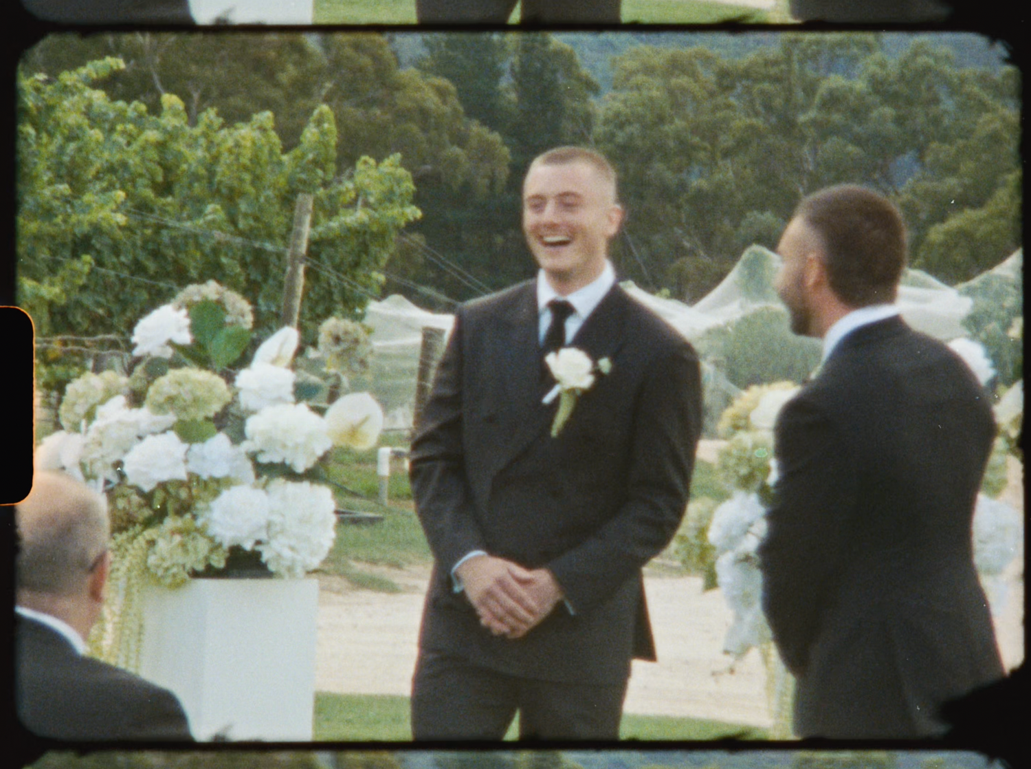 Smiling groom in dark suit with white boutonniere standing at outdoor vineyard wedding ceremony.