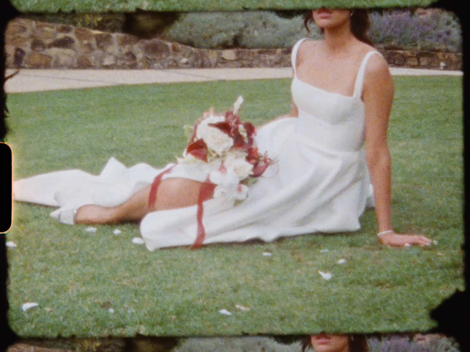 Bride in white dress reclines on grass holding a red and white bouquet.