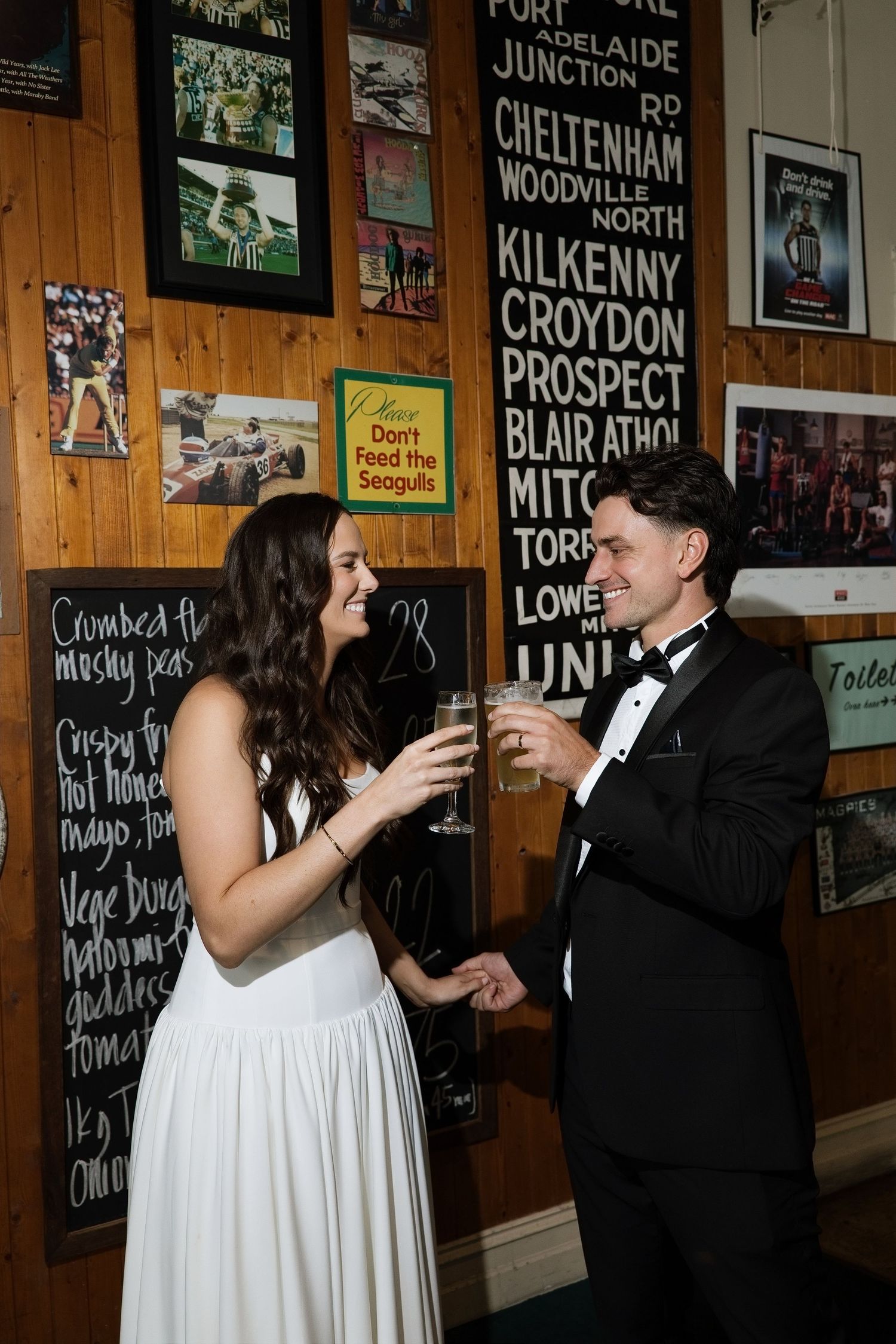 Bride and groom toasting champagne glasses inside a rustic bar with wooden walls and vintage signs.