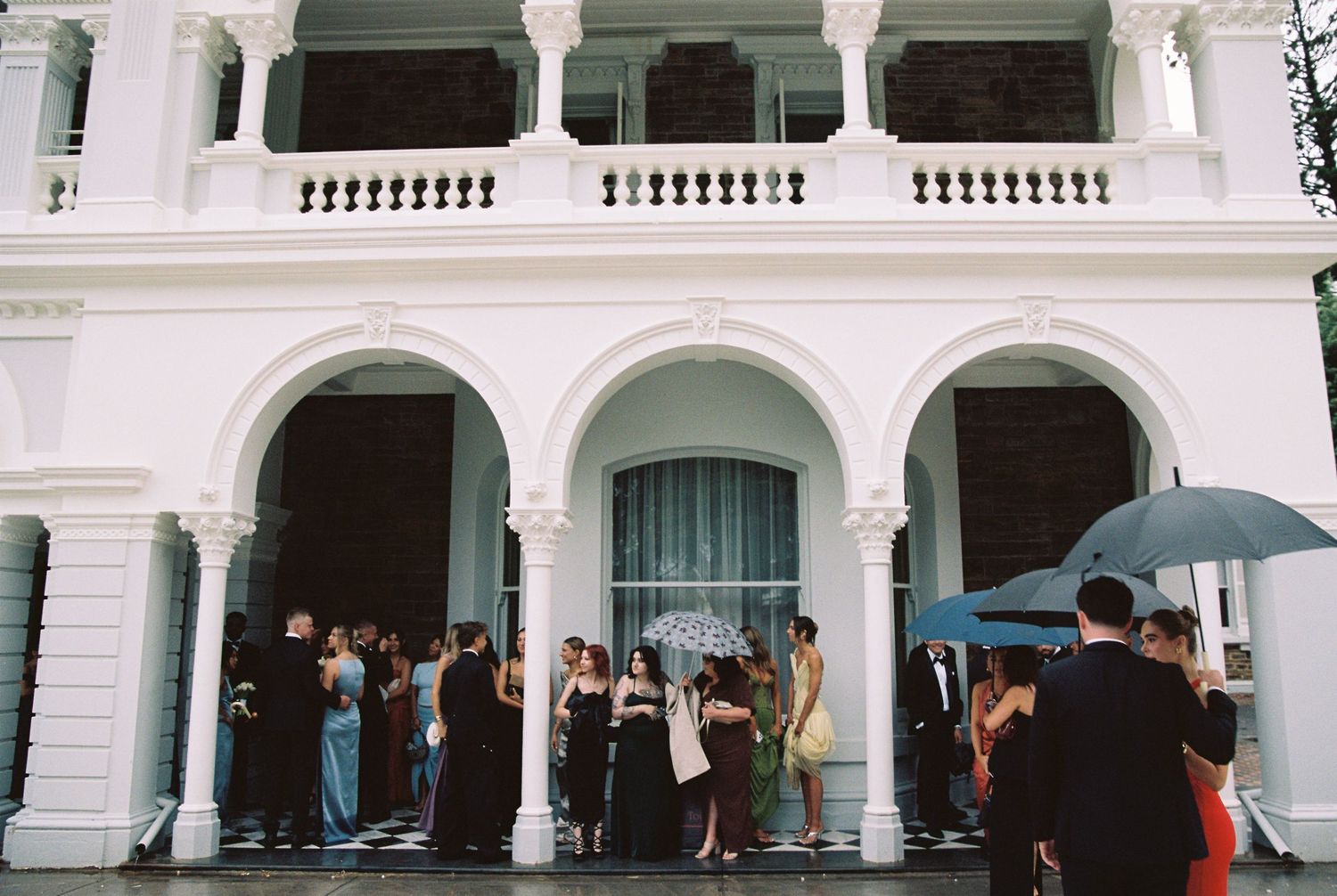 Wedding guests in formal attire gather under umbrellas outside a grand white colonial venue.