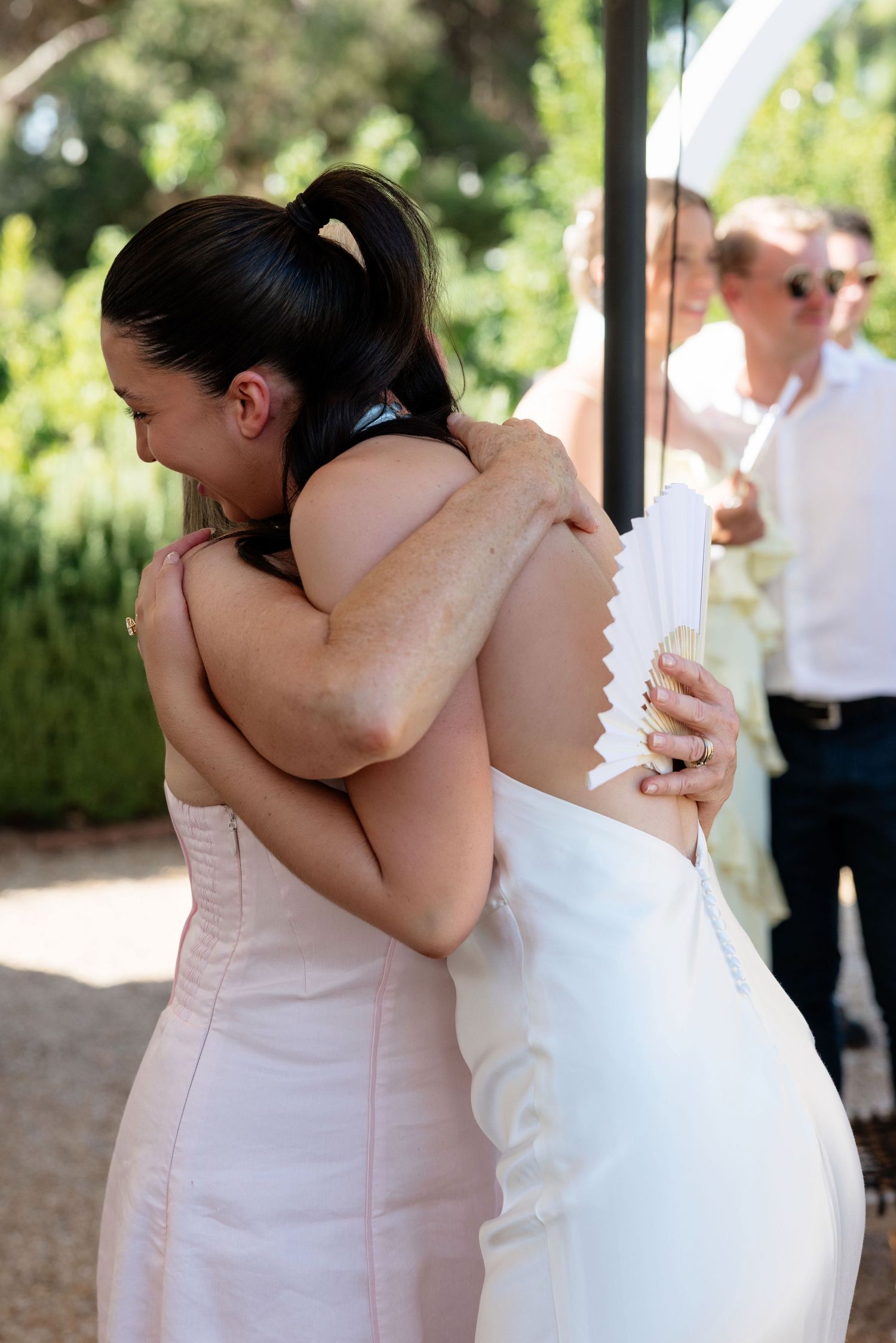 Bride in white gown embracing bridesmaid in pink dress after outdoor wedding ceremony.