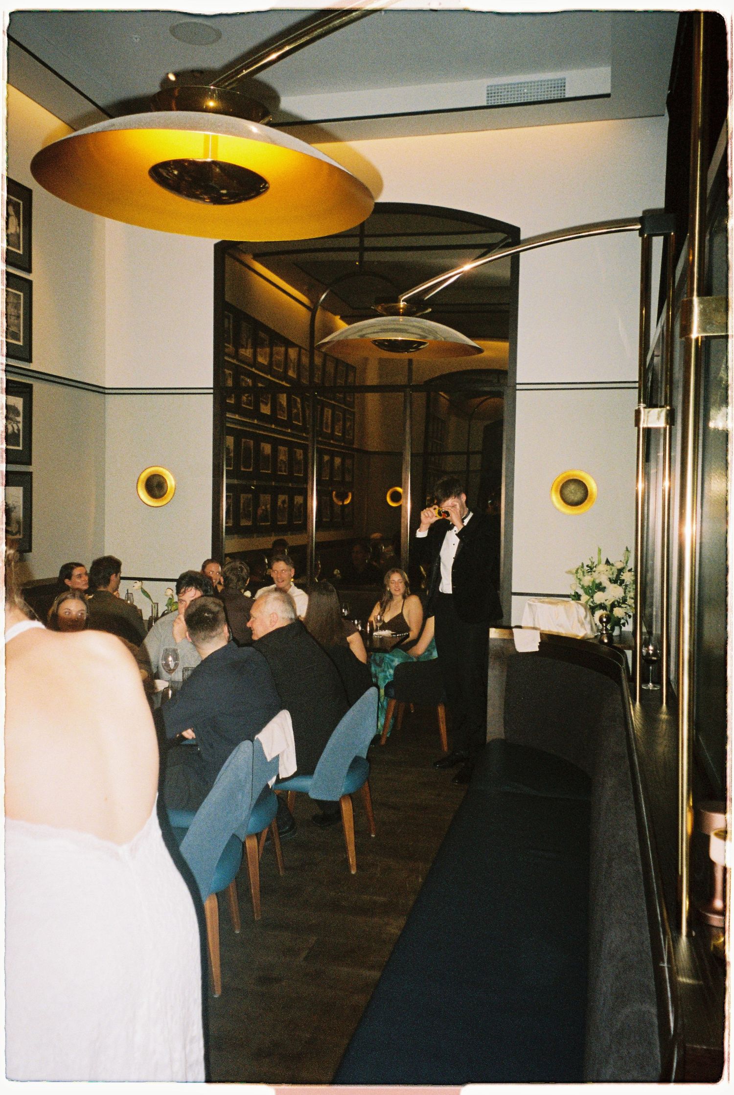 Intimate wedding reception dinner in stylish restaurant with guests seated under gold pendant lights.