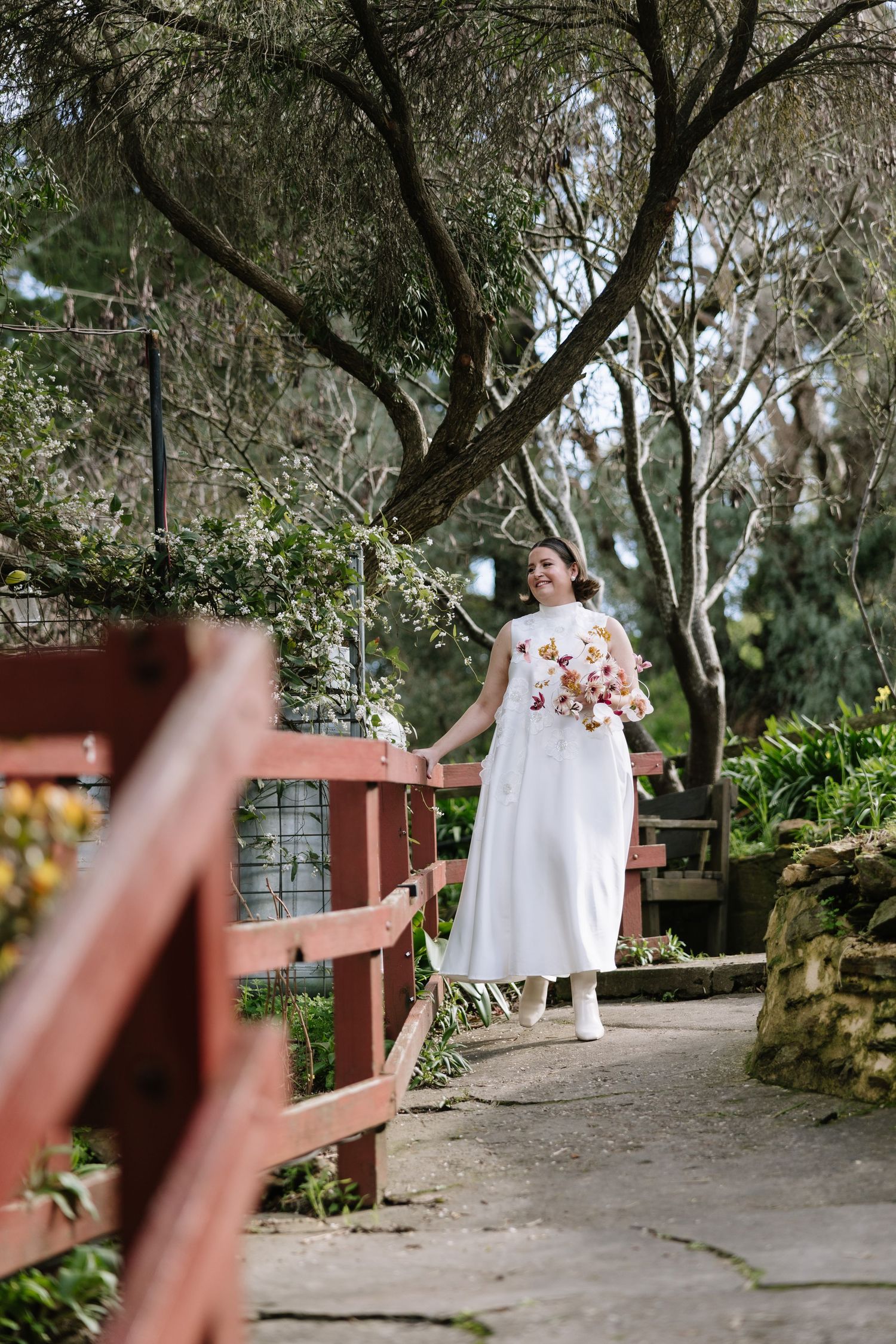 Bride in white gown holding floral bouquet stands on stone path by red bridge surrounded by lush greenery.