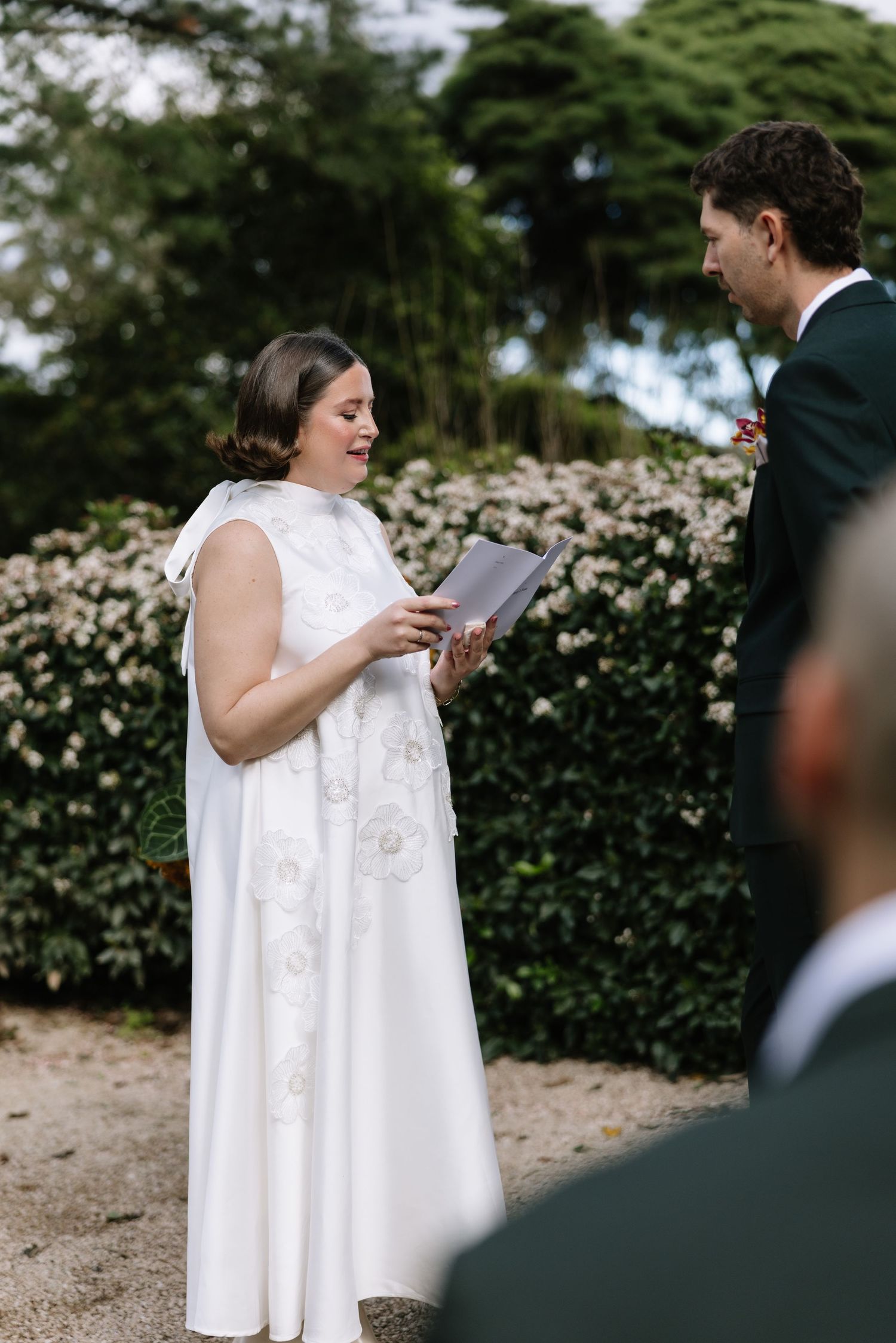 Bride in white gown reads vows to groom outdoors, with flowering hedge backdrop at elegant garden wedding ceremony.