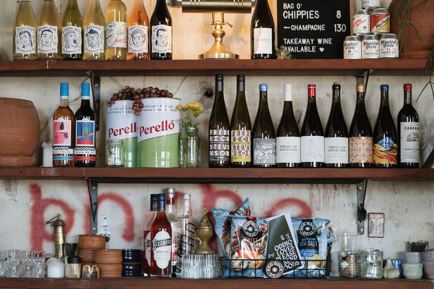 Rustic bar shelves displaying wine bottles, Perello tins, and spirits with a chalkboard menu in a cozy restaurant setting.