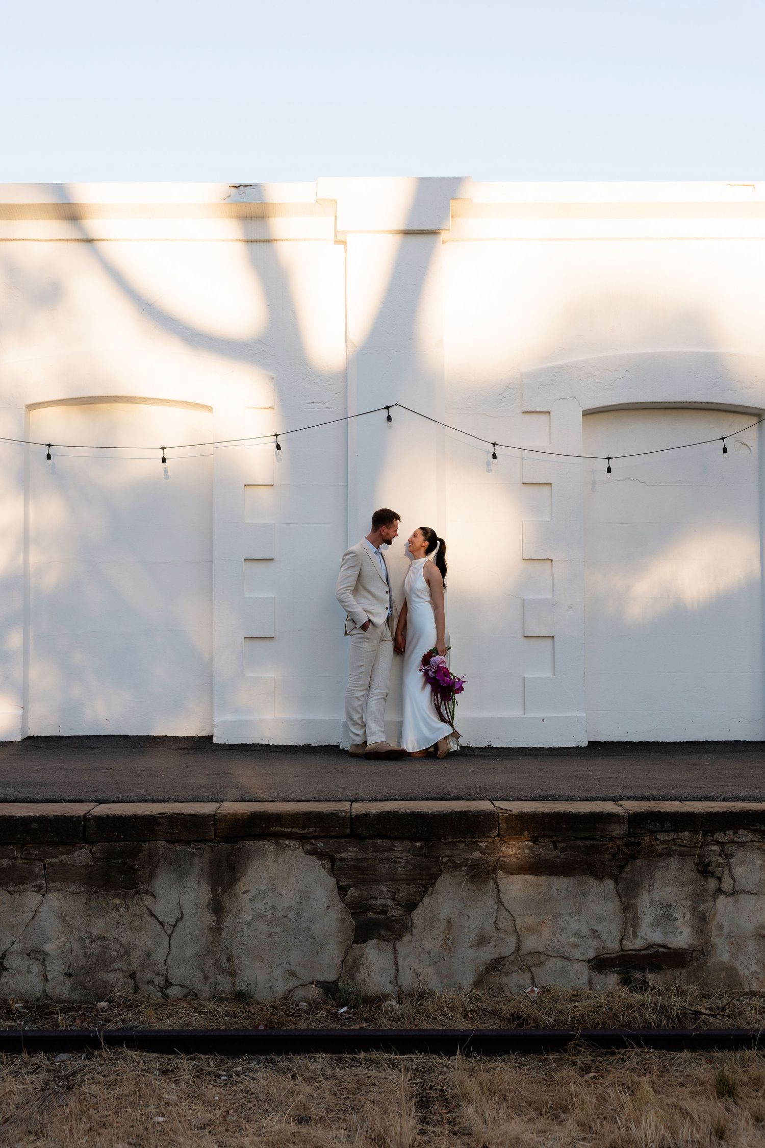 Wedding couple sharing a kiss against white wall with tree shadow at golden hour.