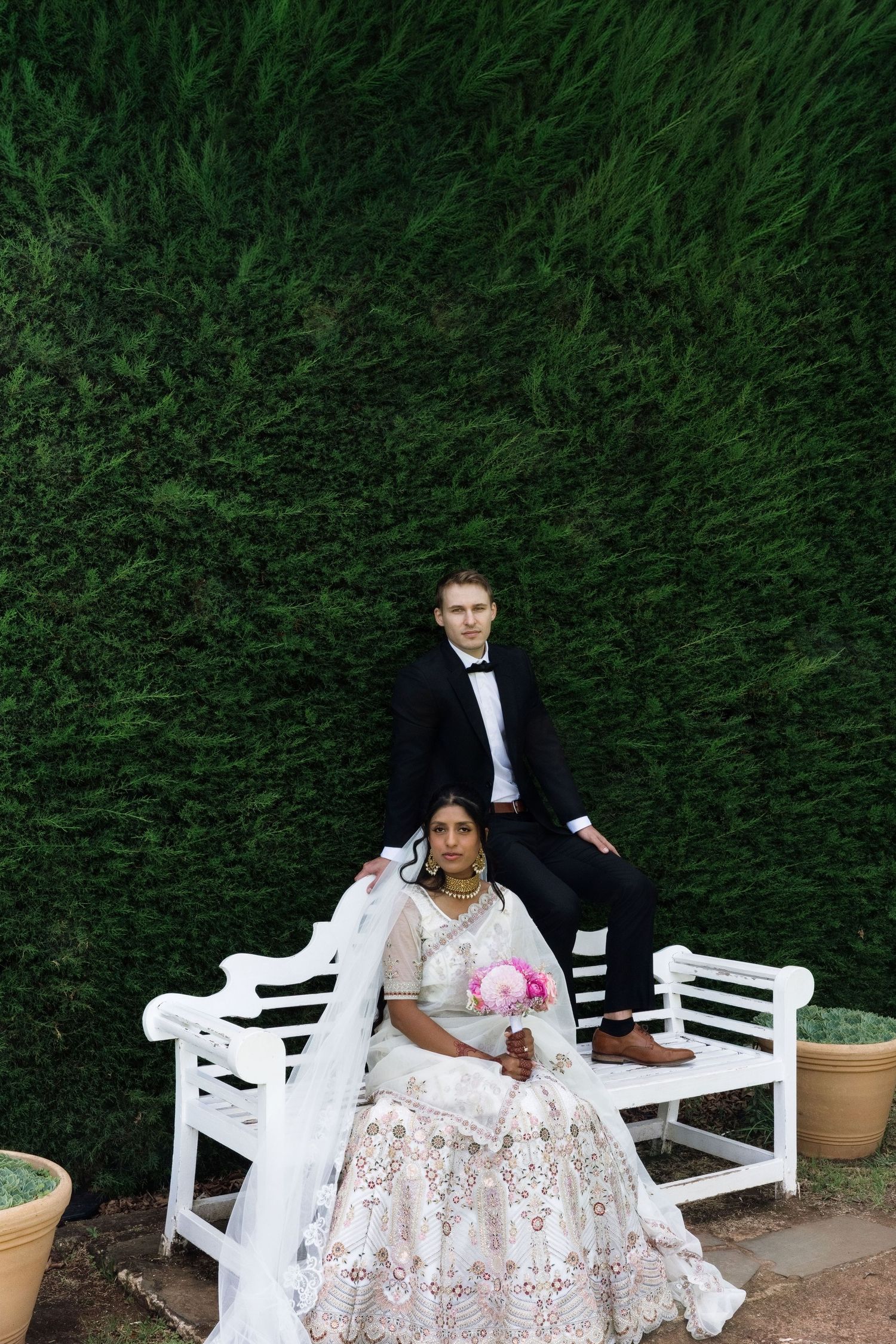 Bride in embroidered lehenga seated on white bench, groom in tuxedo behind her, green hedge background.