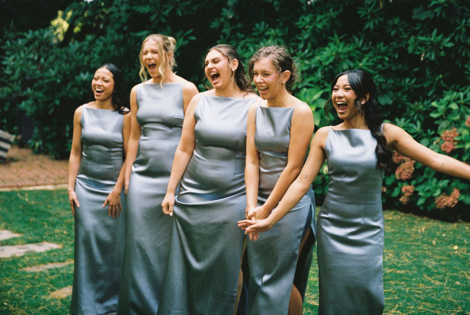 Five bridesmaids in silver satin sleeveless gowns laughing joyfully together outdoors in garden setting.