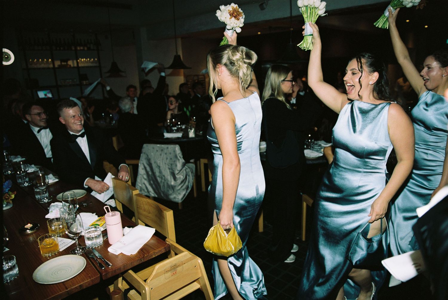 Bridesmaids in blue satin dresses dancing with bouquets at wedding reception.