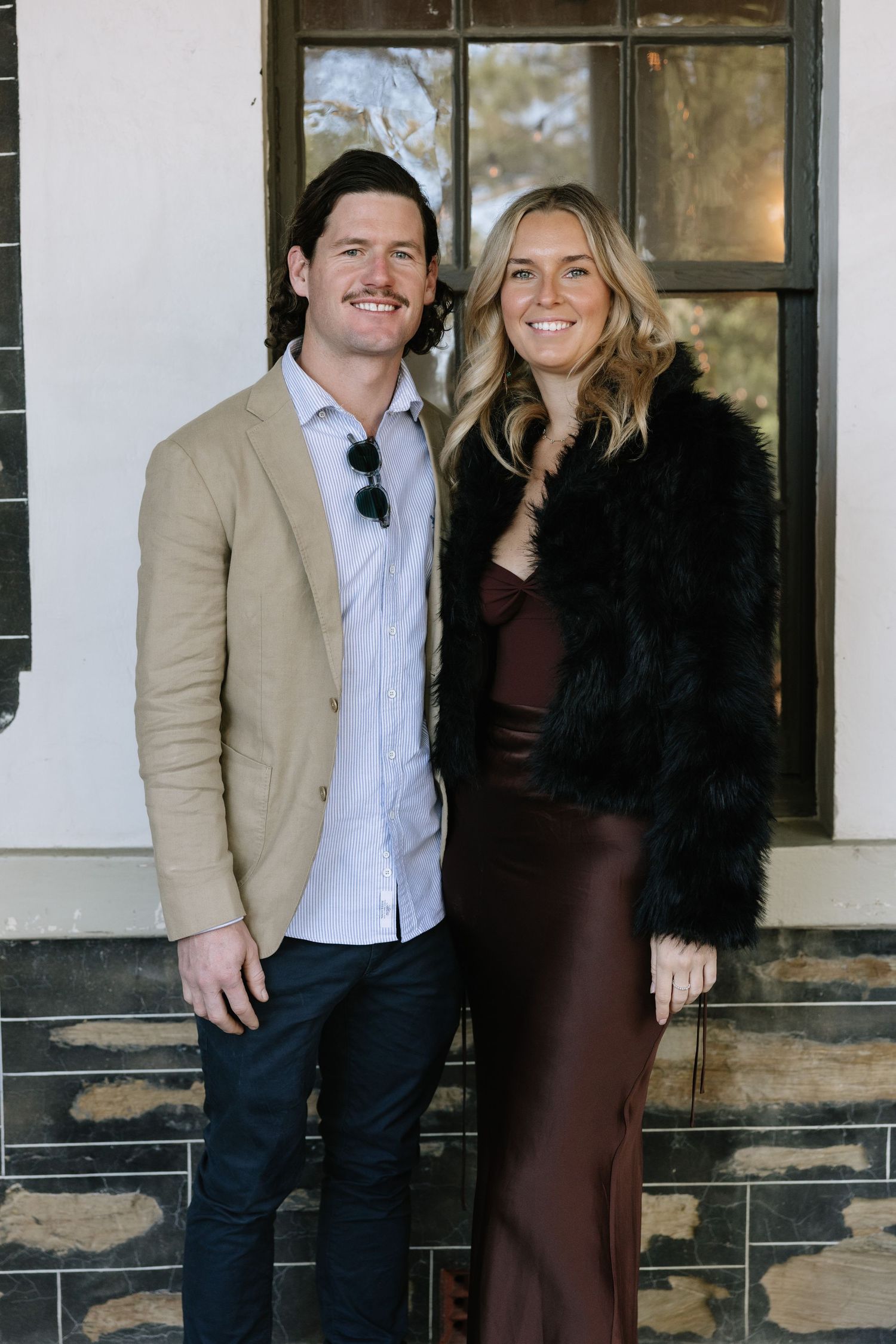 A smiling couple poses outdoors, man in tan blazer and woman in burgundy satin dress with black fur coat.
