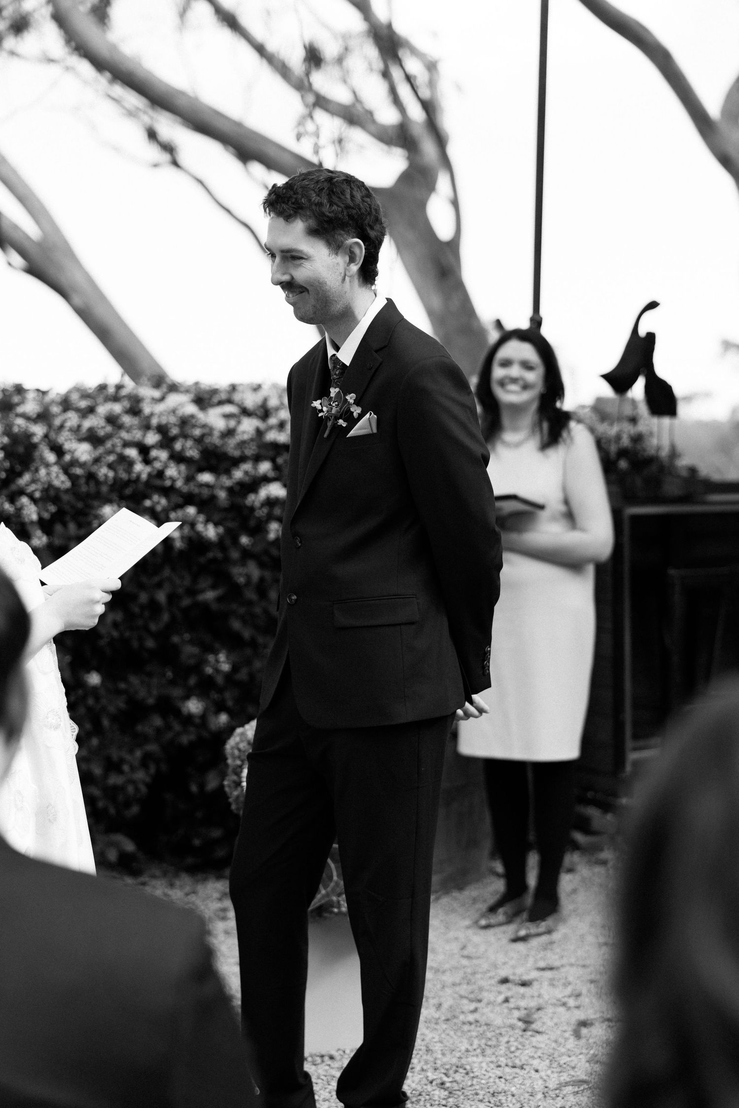 Groom smiling during outdoor wedding ceremony in suit with boutonniere, black and white photo.