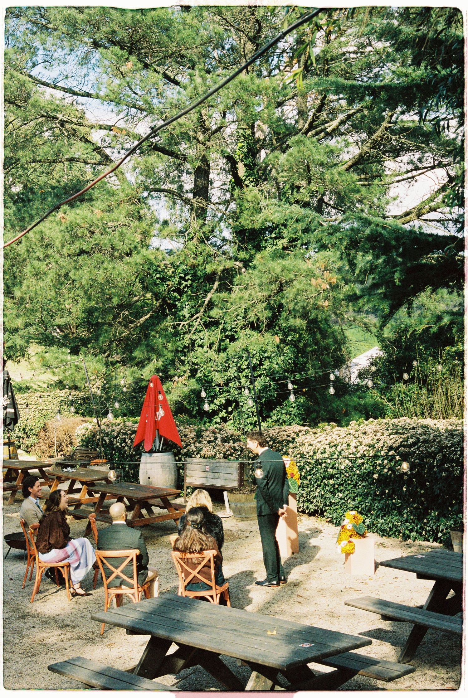 Outdoor garden patio with picnic tables, red umbrella, lush green trees, and people dining al fresco on a sunny day.