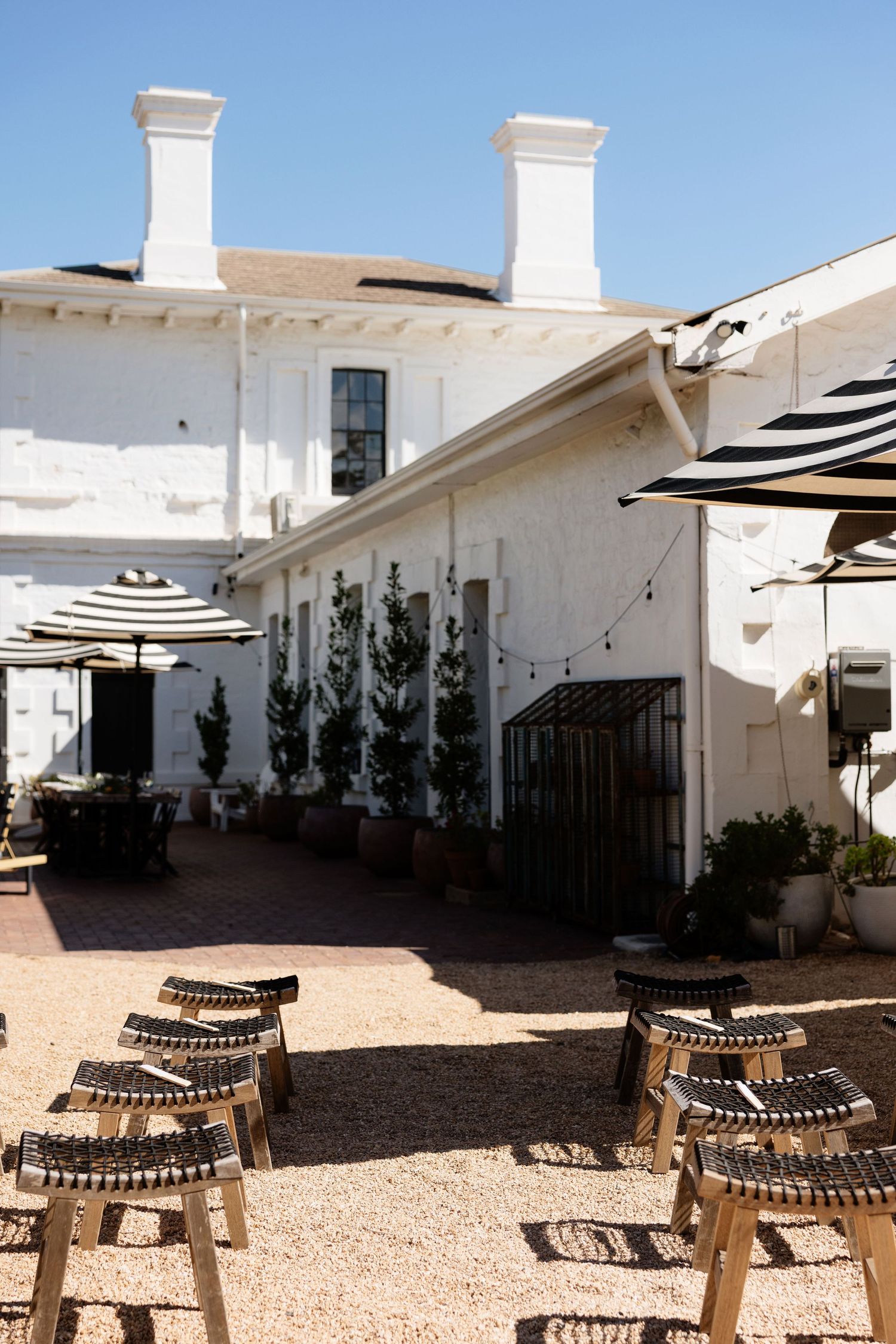 Charming white-washed outdoor courtyard with woven stools, striped umbrellas, potted trees, and string lights on a sunny day.