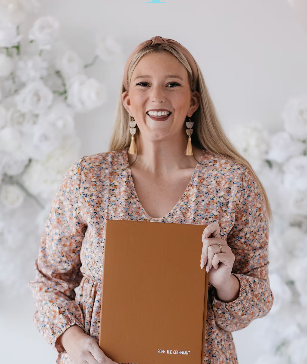 Smiling woman in floral dress holding a tan leather folder in front of white flower backdrop.