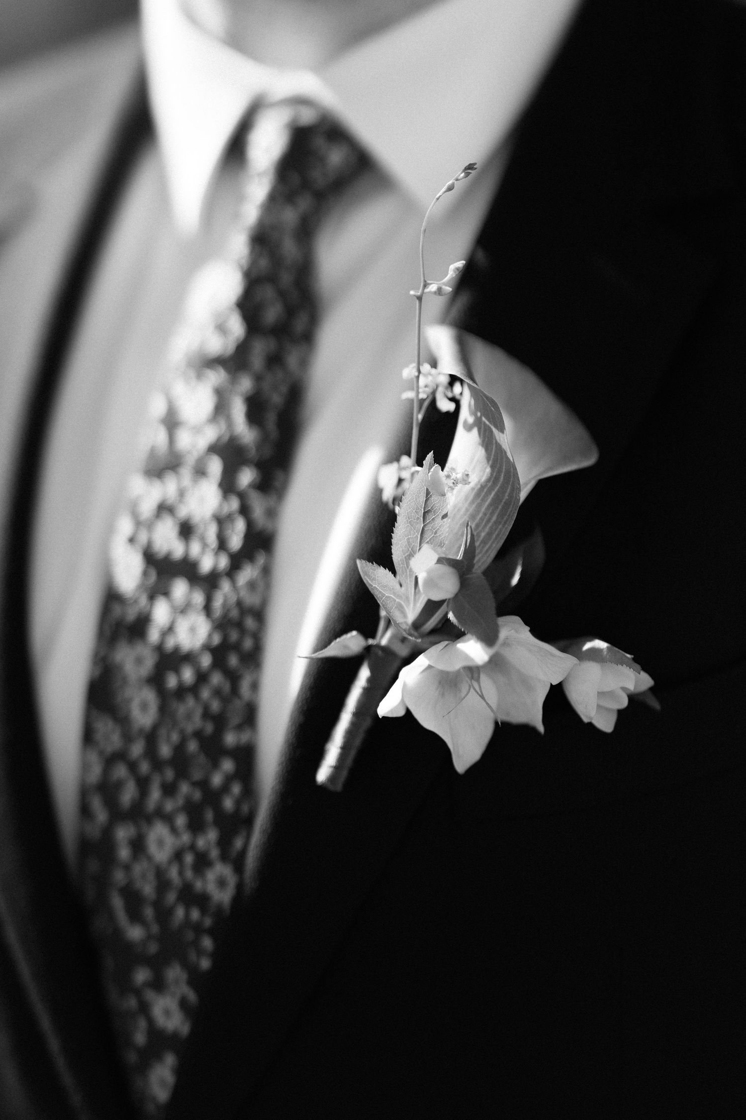 Black and white close-up of groom's floral boutonniere and patterned tie on dark suit.