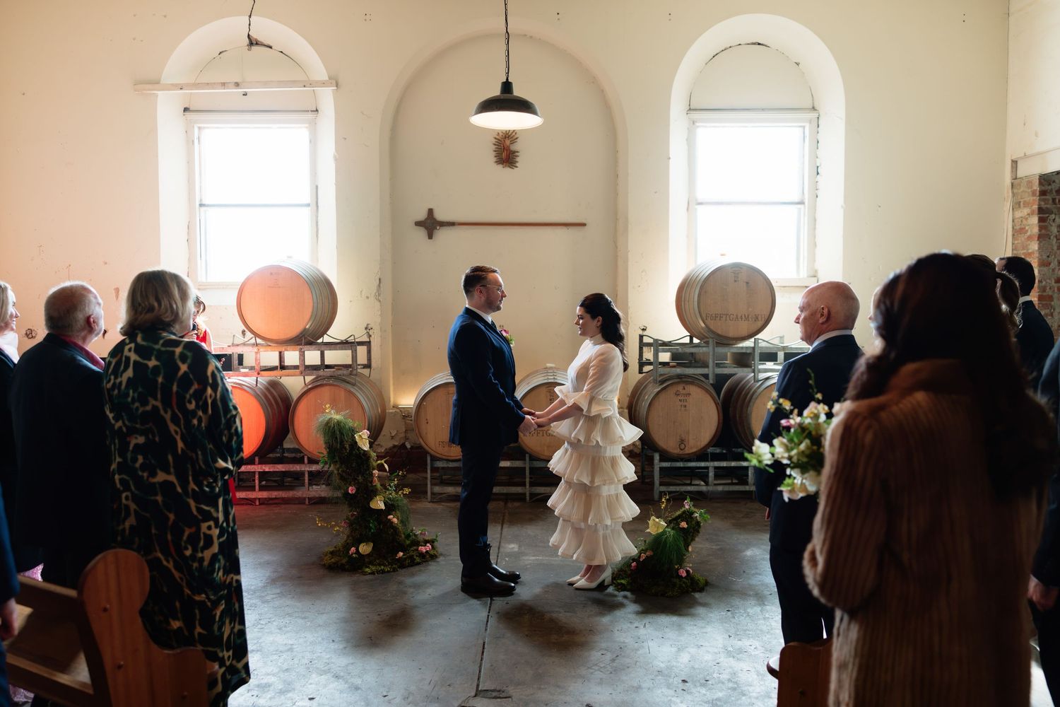 Couple exchanges wedding vows in a rustic winery ceremony with oak barrels and guests gathered around.
