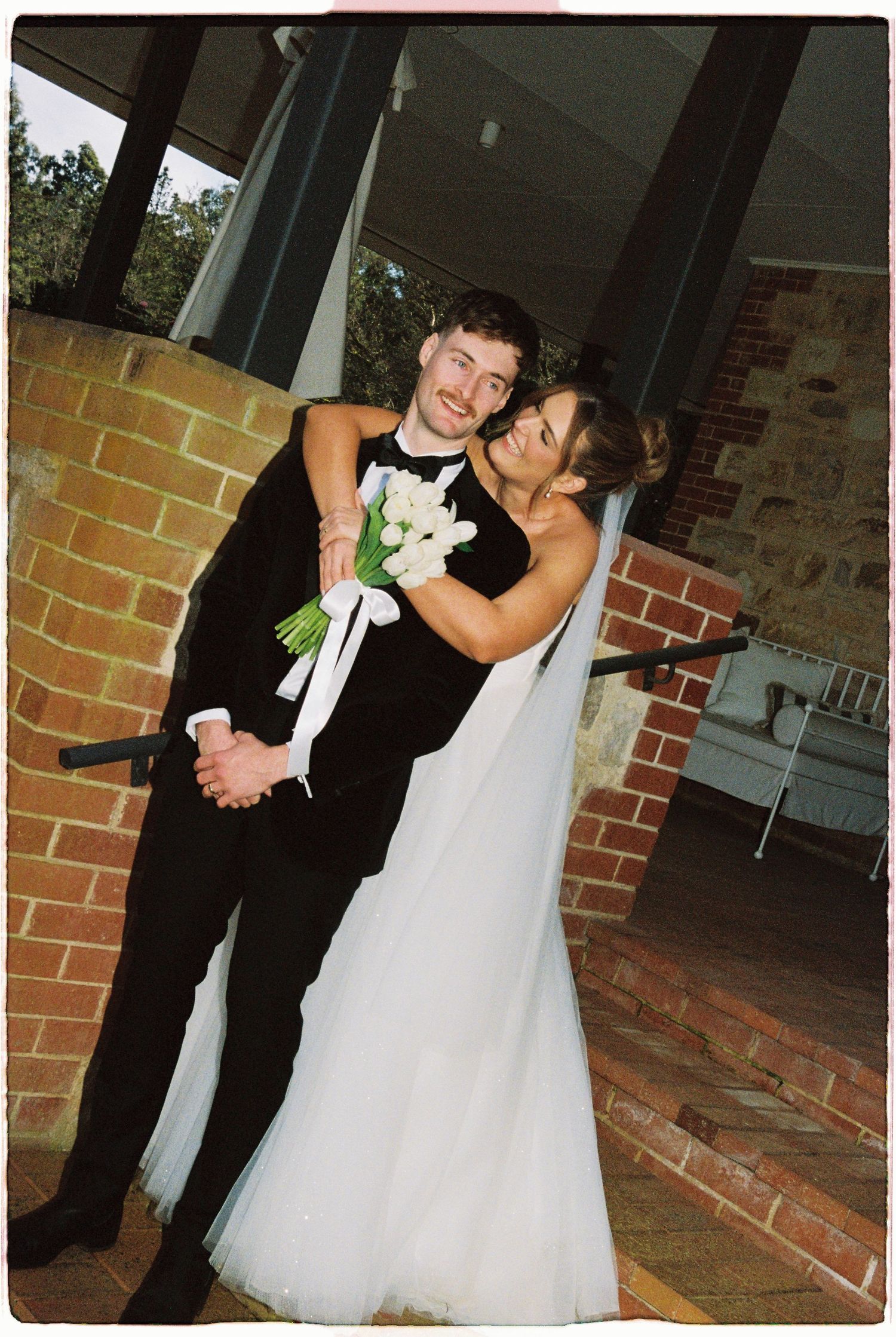Bride and groom embrace near a brick wall, with the bride holding a bouquet of white tulips.