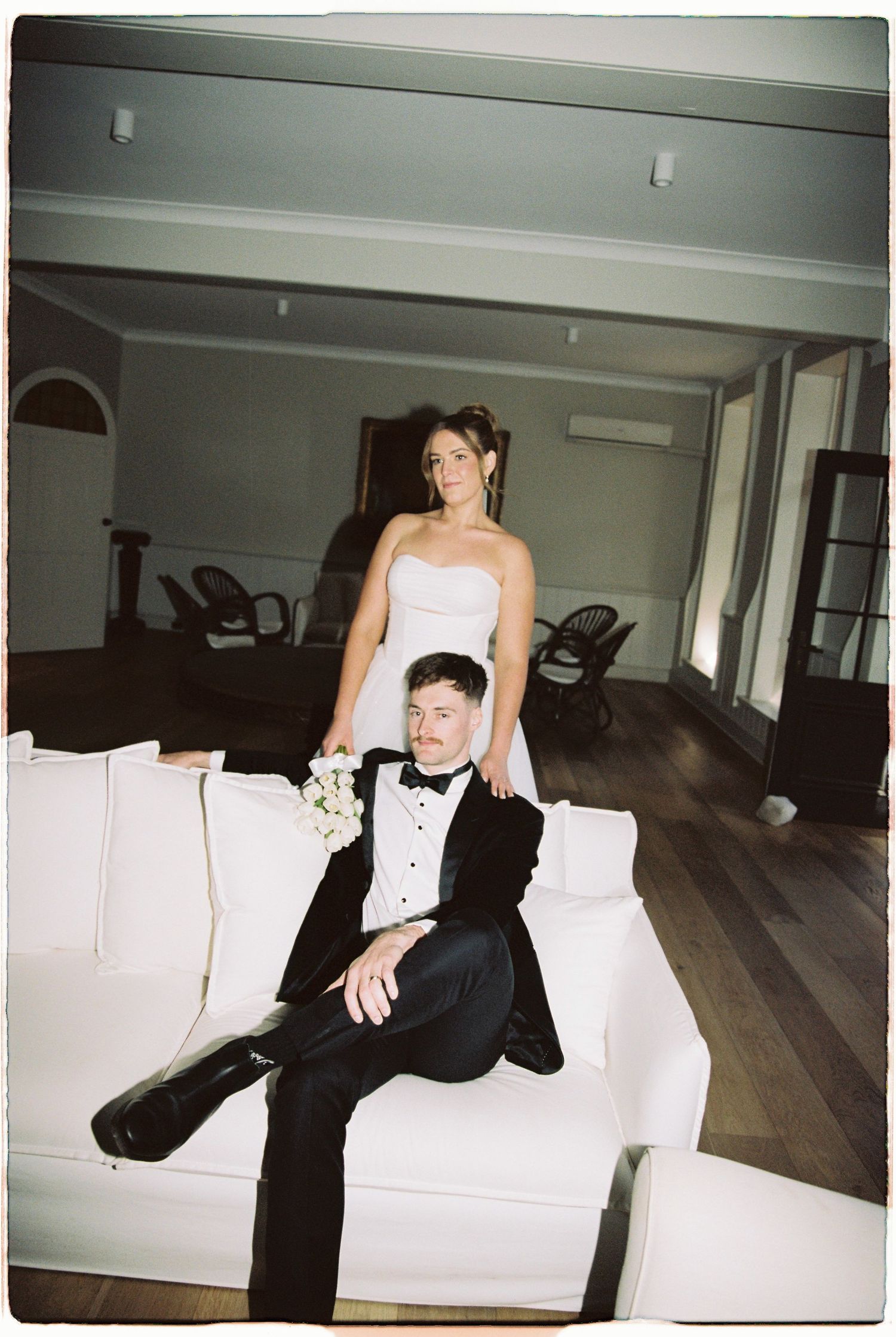 Bride standing behind groom seated on white sofa in elegant wood-floored reception room.