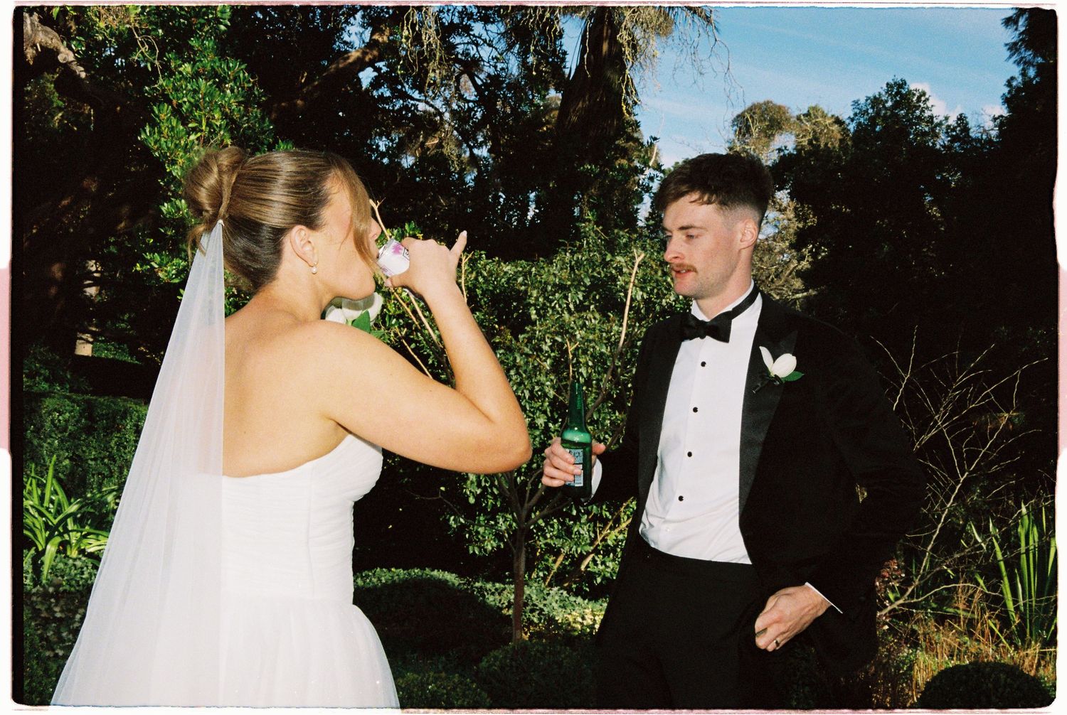 Bride in white gown and veil drinks from bottle while groom in black tuxedo watches at outdoor wedding.