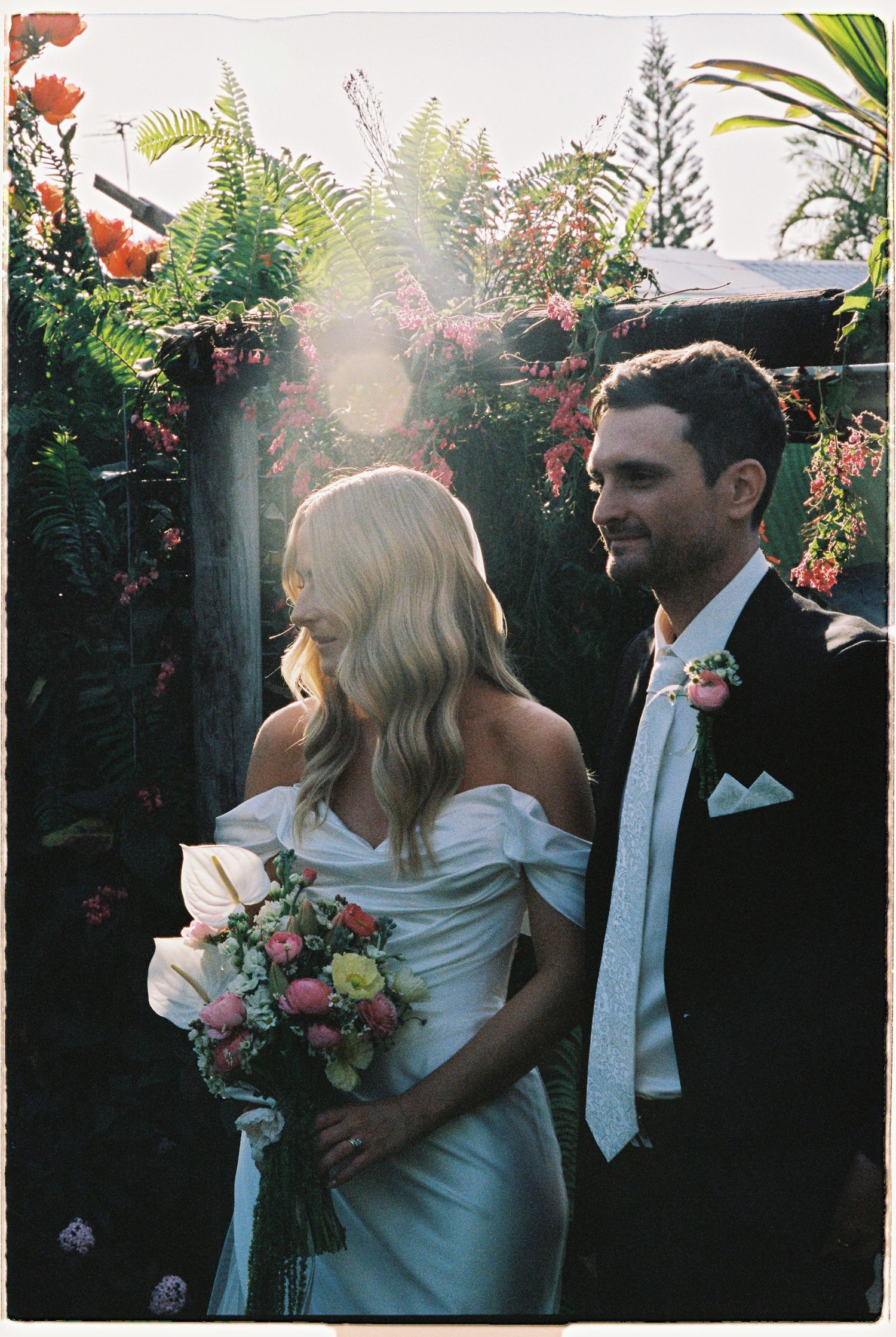 Bride and groom share a romantic moment surrounded by lush garden greenery and sunlight at outdoor wedding.
