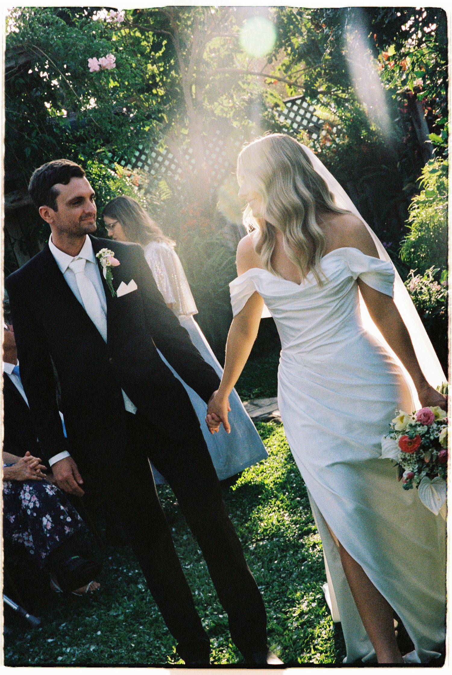 Bride and groom walking hand in hand through sunlit garden after outdoor wedding ceremony.