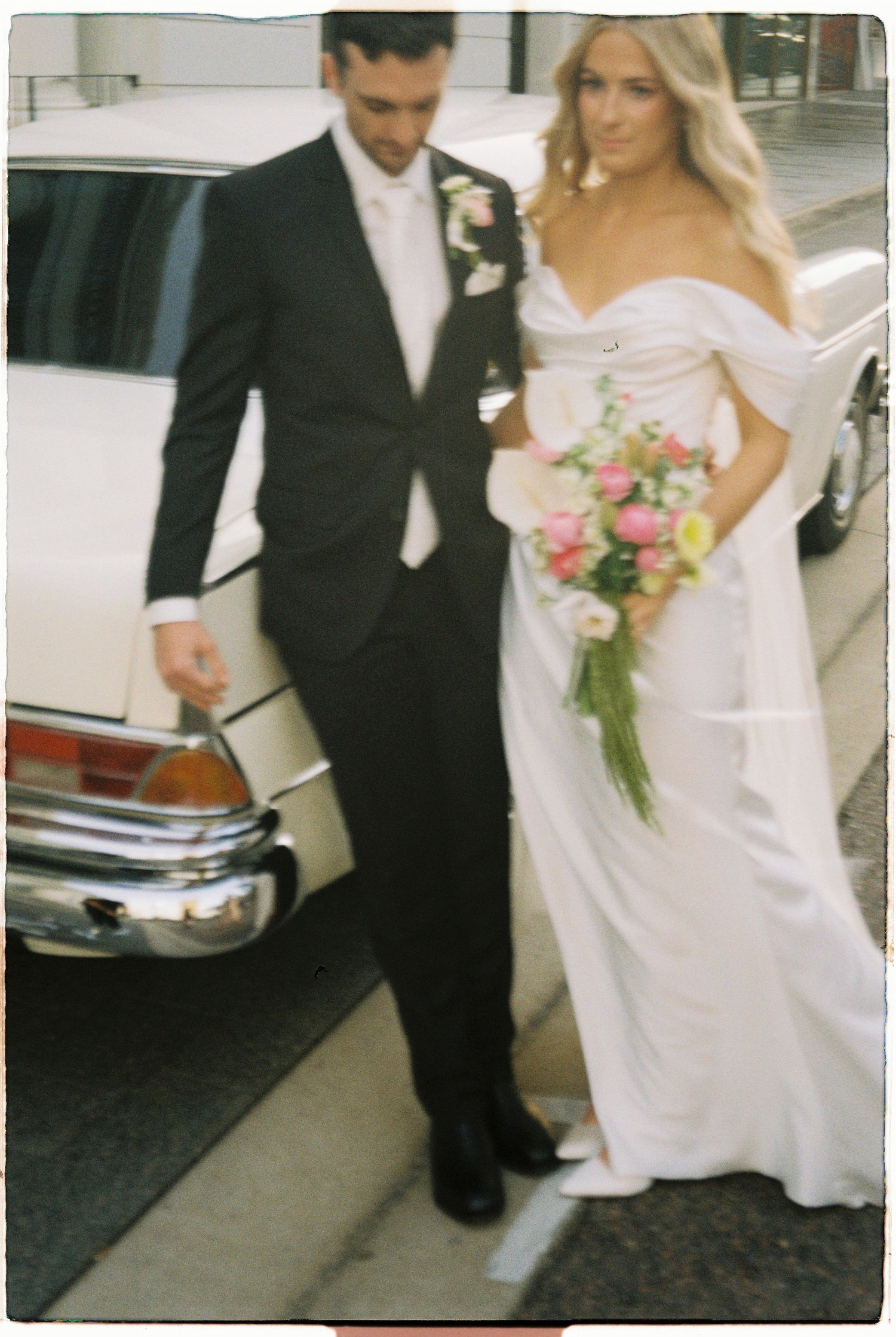 Elegant bride in off-shoulder white gown holds vibrant bouquet beside groom in black suit near a classic white vintage car.