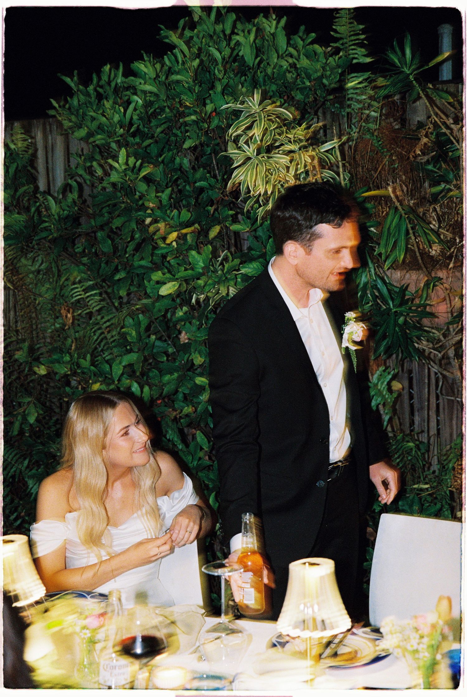 A smiling bride in white off-shoulder gown sits beside a groom in a black suit at a garden wedding reception.