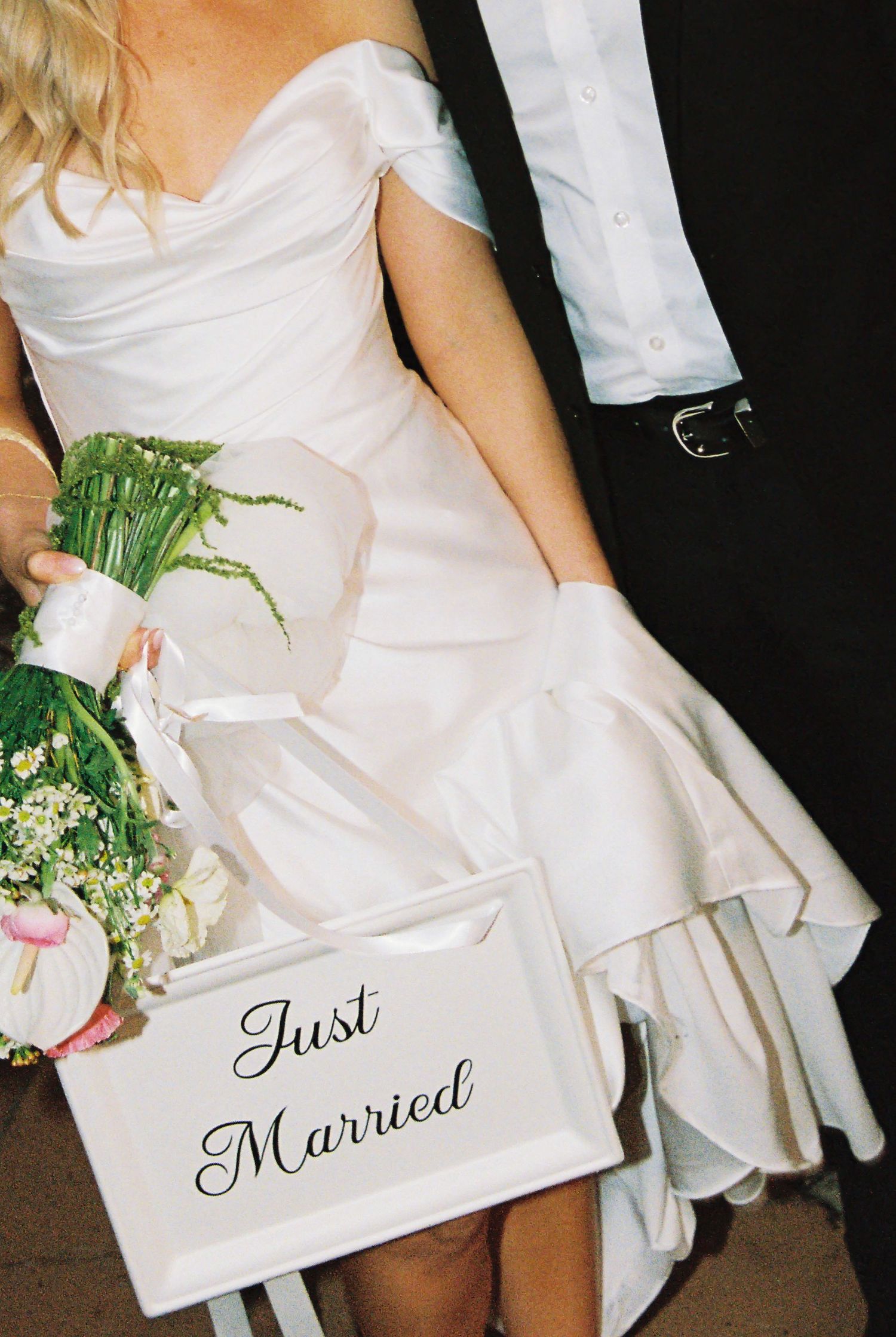Bride in white off-shoulder gown holding bouquet with a Just Married sign at her wedding.