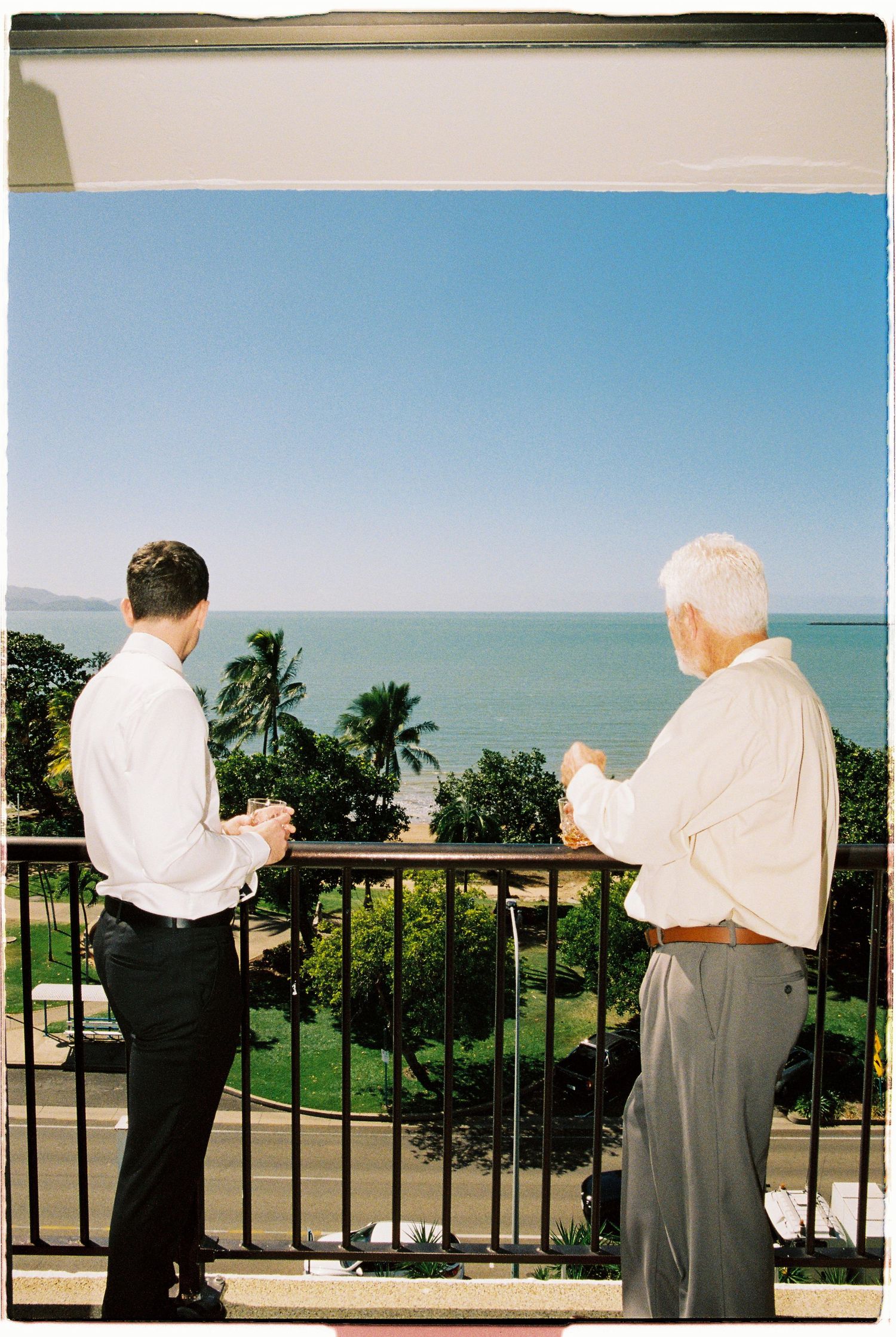 Two men in white shirts enjoy drinks on a tropical balcony overlooking lush palm trees and a calm blue ocean.