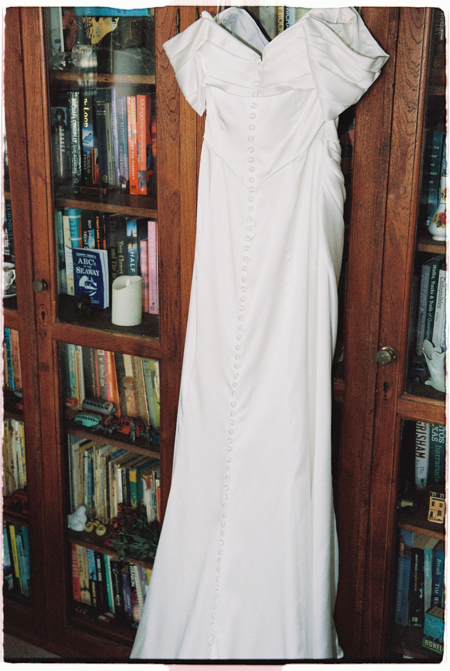 White off-shoulder wedding gown with button back detail hanging in front of a wooden bookshelf.