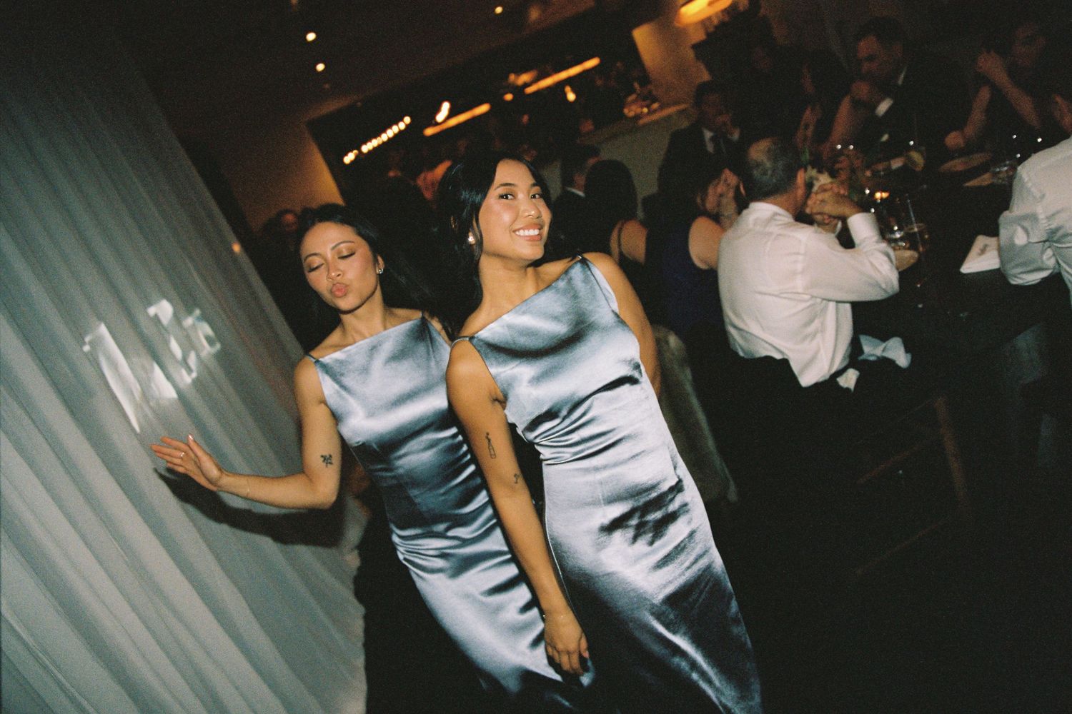 Two bridesmaids in silver satin dresses smile and dance at an elegant wedding reception.