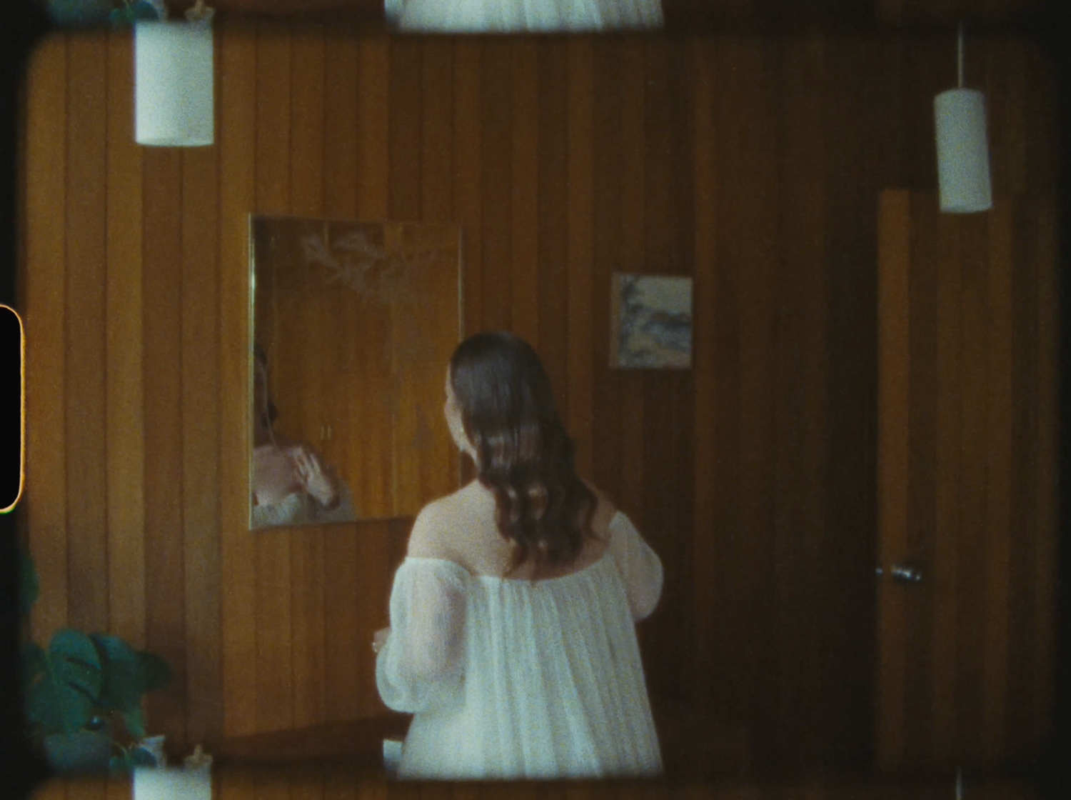 A woman in a white dress stands in a wood-paneled room near a mirror.