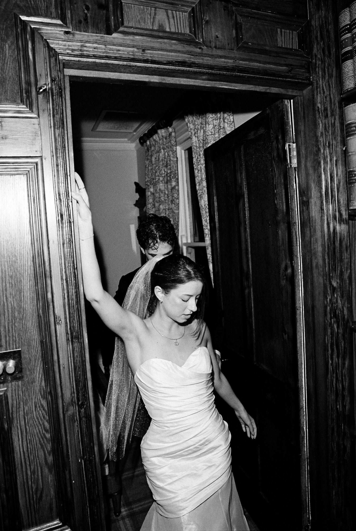 Black and white photo of a bride in a strapless wedding gown and veil walking through a wooden doorway.