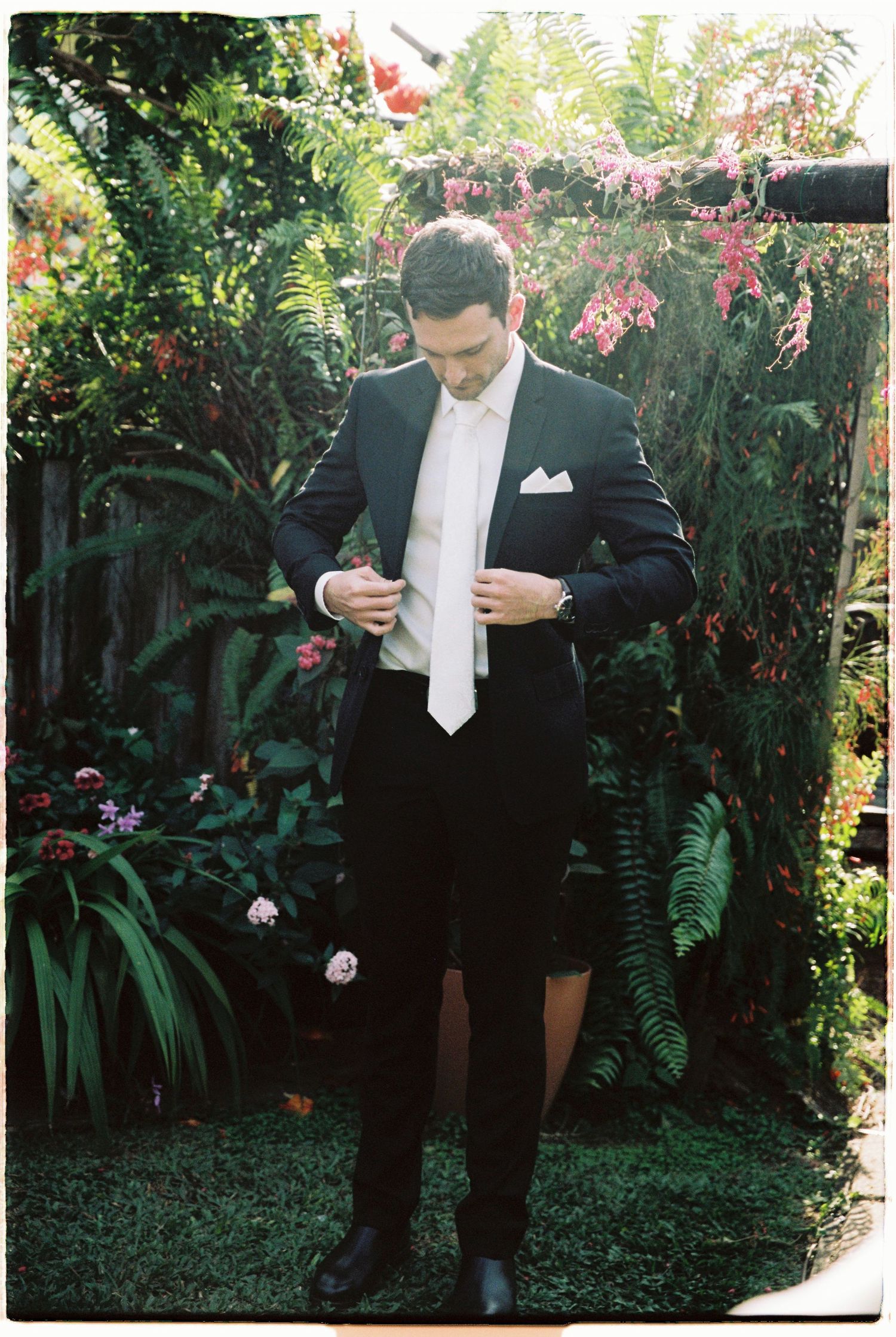 Groom in black tuxedo adjusting his jacket in a lush tropical garden with vibrant flowers.