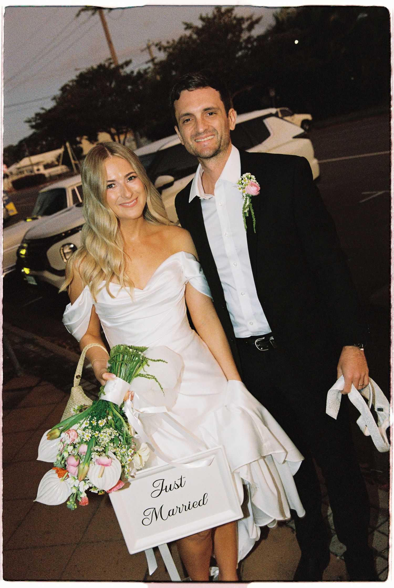 Newlywed couple smiling outdoors holding a Just Married sign, bride in off-shoulder gown with bouquet.