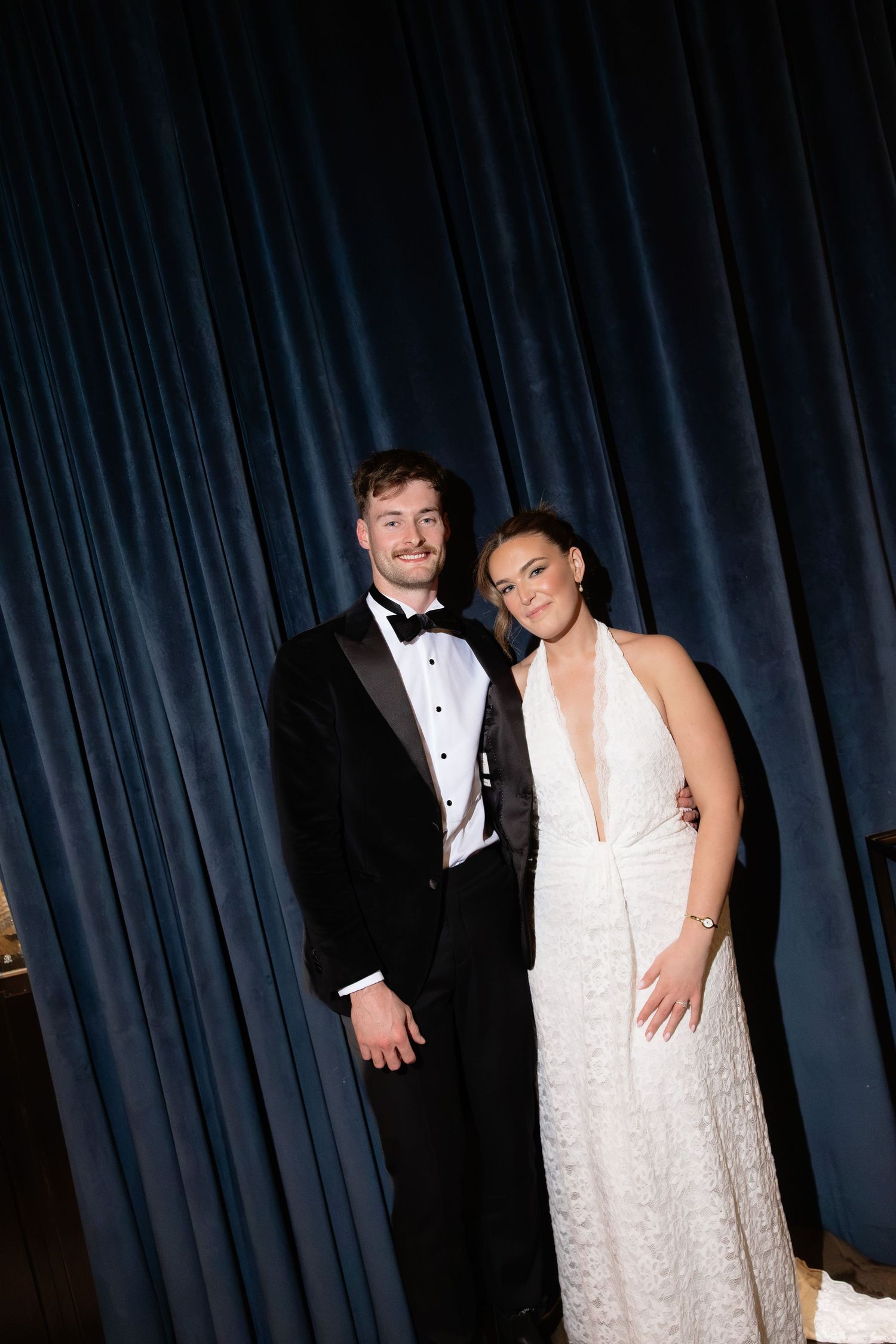 A couple in formal attire, man in black tuxedo and woman in white halter gown, posing before navy blue velvet curtains.