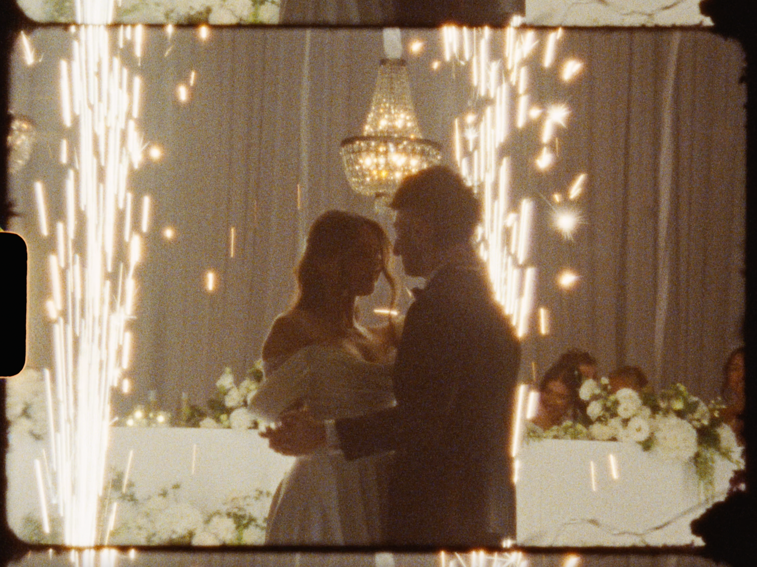 Couple sharing first dance at wedding reception with sparkling fireworks backdrop and elegant chandelier lighting.