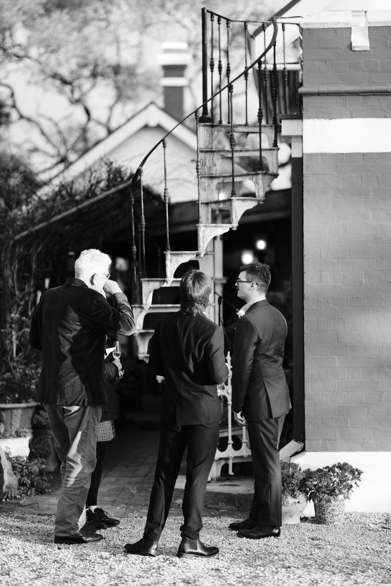 Black and white photo of elegantly dressed guests conversing outside a venue with a spiral staircase.
