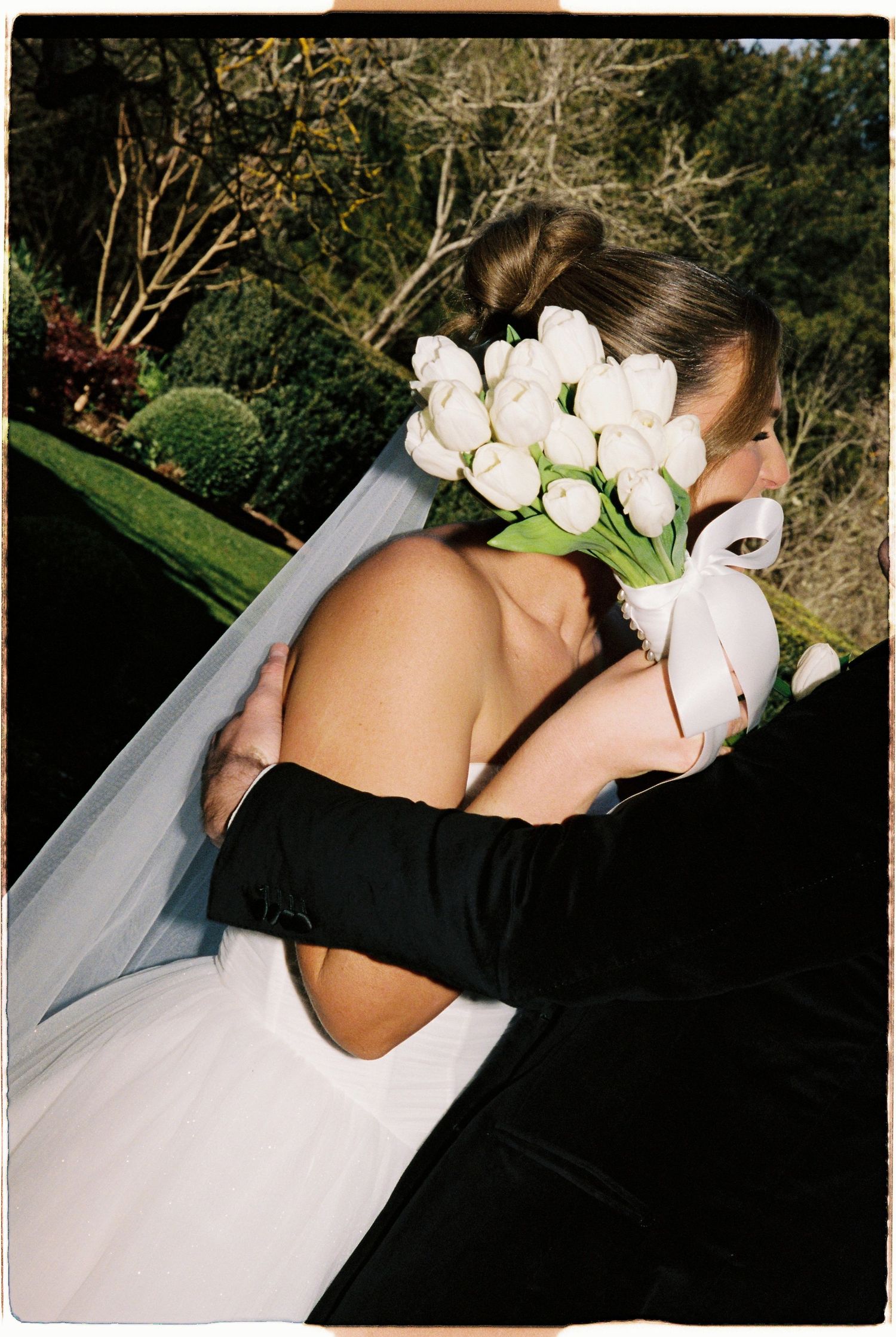 Bride in backless white gown holding white tulip bouquet embraced by groom in black suit outdoors.