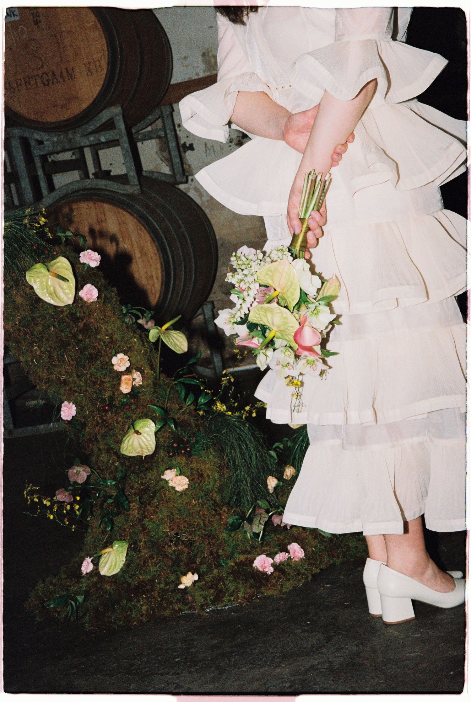 Bride in white ruffled dress holding a pastel bouquet beside moss and pink flowers near rustic wooden barrels.